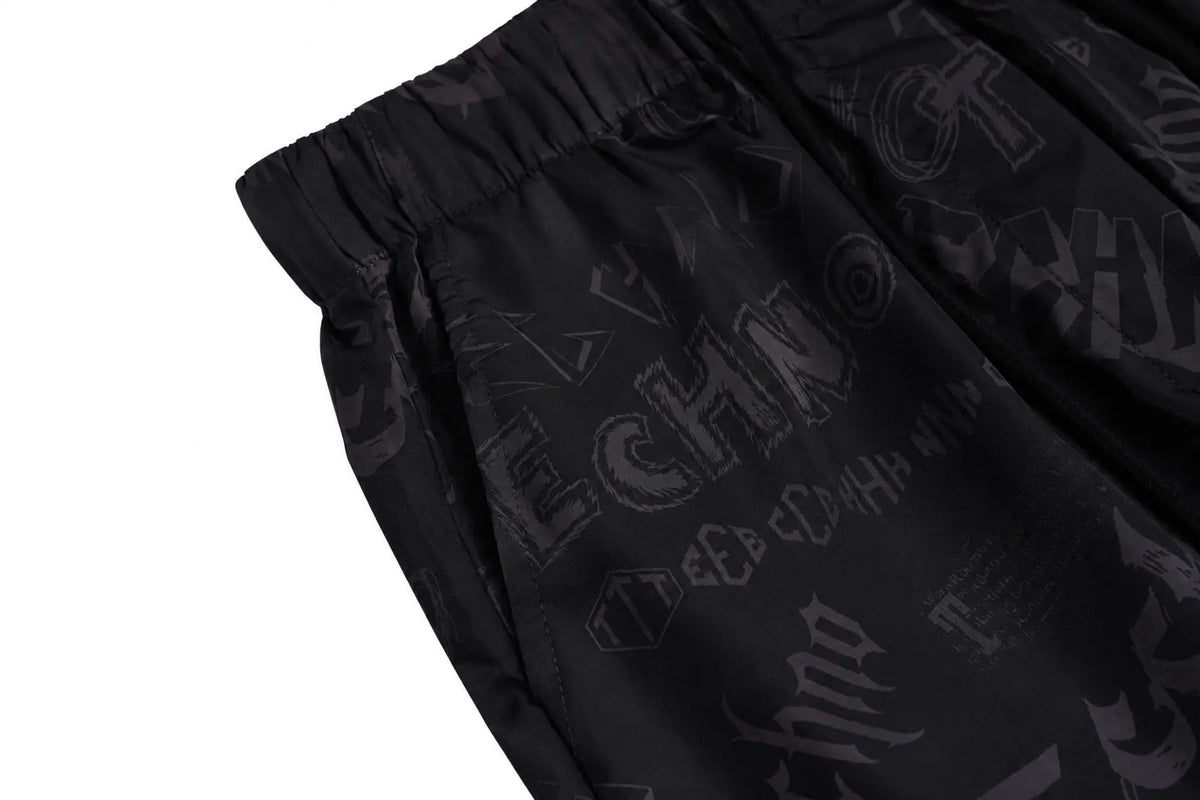 Close-up of black athletic shorts with an elastic waistband and a tonal dark gray graphic print featuring the word 'ECHO' and other stylized text, shown on a white background. TCH Techno Logo Wide-Leg Cargo Pants