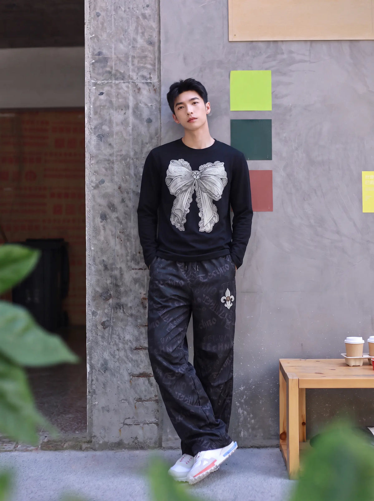 A man stands in a concrete-walled indoor setting wearing a black long-sleeve shirt with a large gray bow graphic on the front, dark patterned pants, and white sneakers. TCH Techno Logo Wide-Leg Cargo Pants