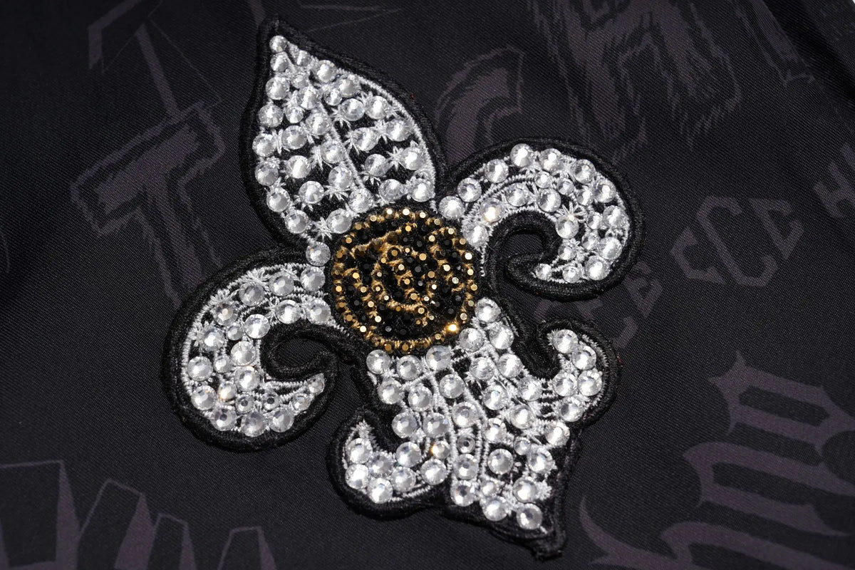 A close-up of a fleur-de-lis patch embellished with clear rhinestones and a gold rose center, applied to a black garment with a tonal gothic font pattern. TCH Techno Logo Wide-Leg Cargo Pants