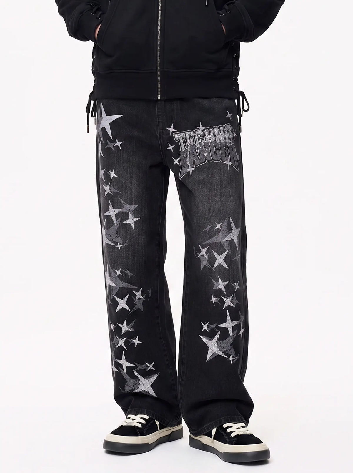 A person wearing black jeans with a distressed wash and scattered silver star graphics, paired with a black zip-up jacket and black sneakers with white laces, shown against a plain white studio background. TCH Techno Magic Star Graphic Wide-Leg Jeans