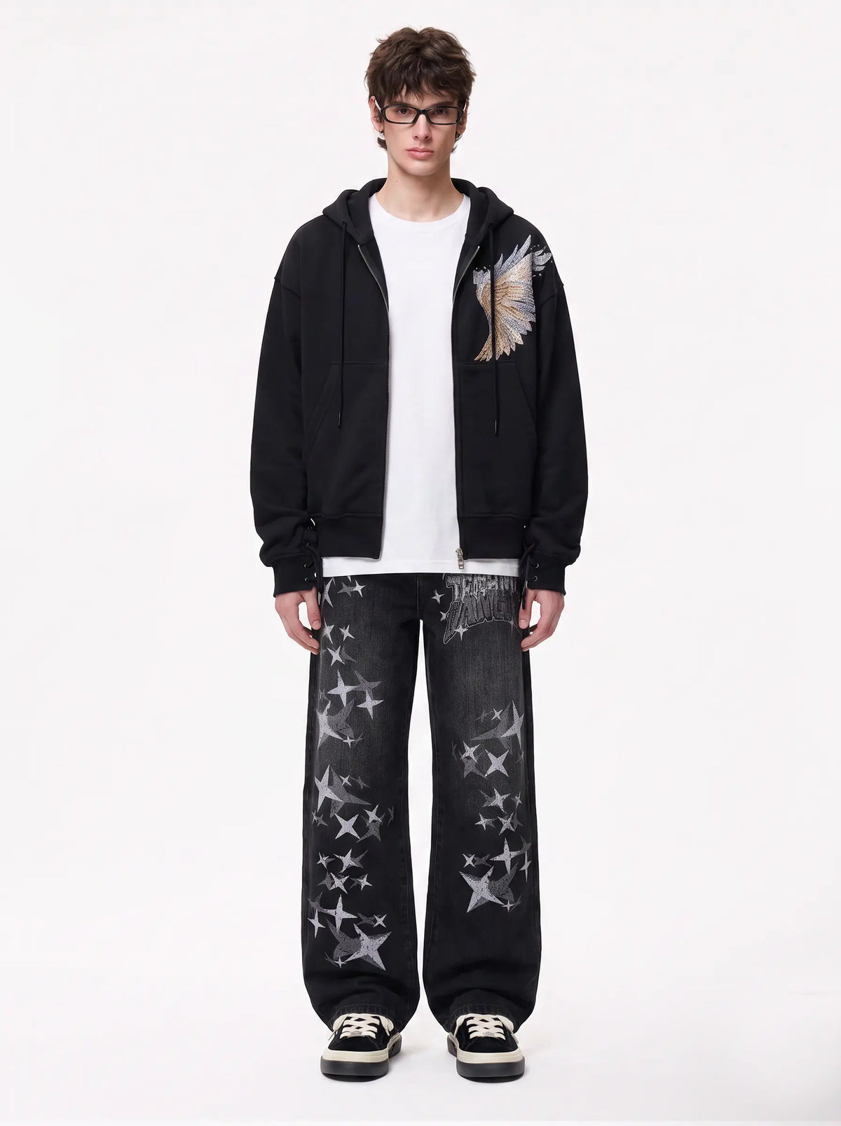 A model stands against a plain white studio background wearing a black zip-up hoodie with a large wing graphic on the left chest, a white t-shirt, black jeans with white star prints, and black-and-white sneakers. TCH Techno Magic Star Graphic Wide-Leg Jeans