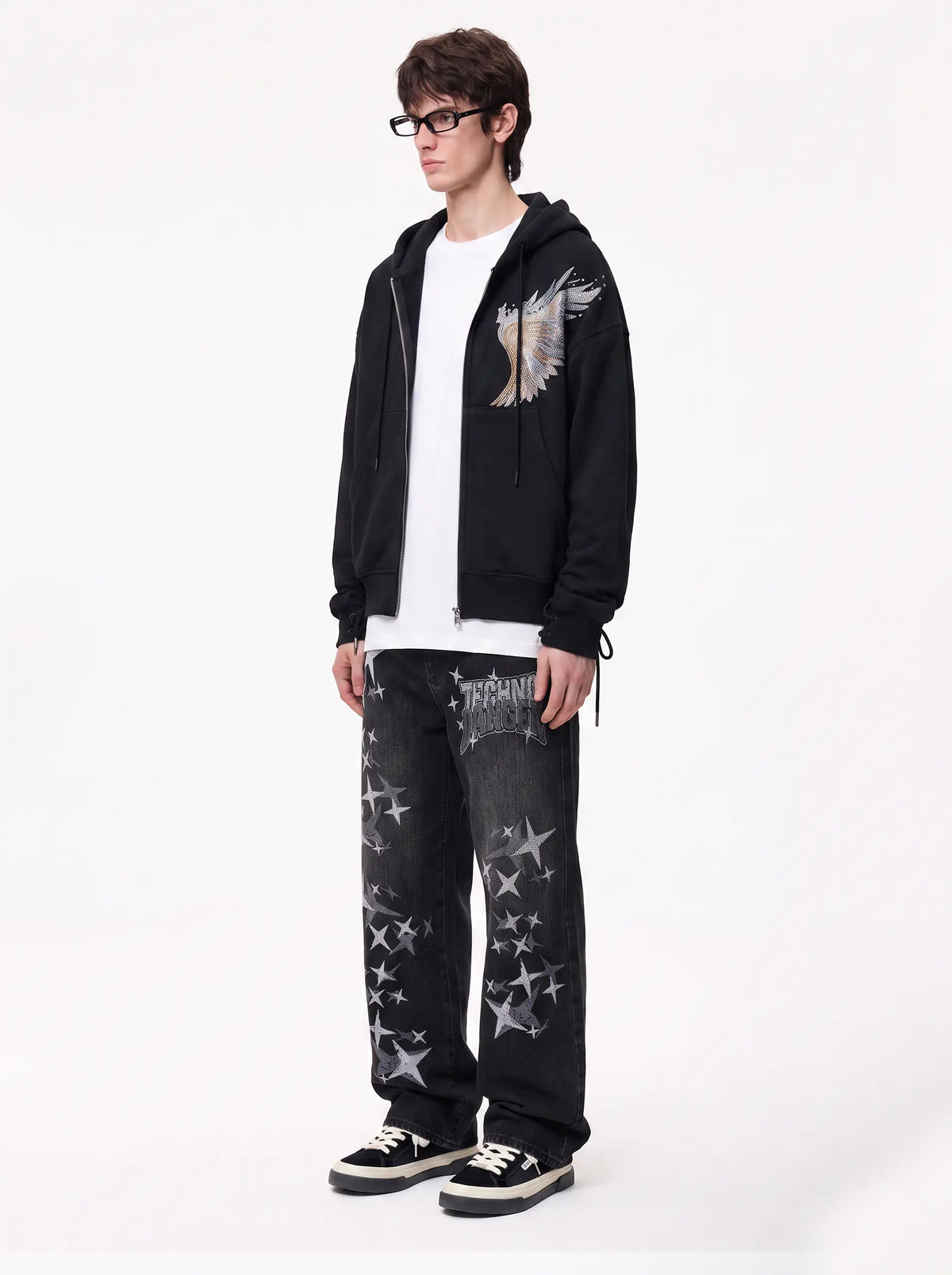 A model wearing a black zip-up hoodie with a wing graphic on the chest, a white t-shirt, black jeans with white star prints, and black sneakers, standing against a plain white studio background. TCH Techno Magic Star Graphic Wide-Leg Jeans