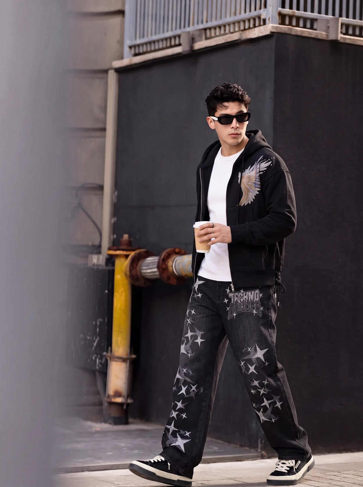 A man walking outdoors wearing a black zip-up hoodie with a wing graphic, a white t-shirt, black jeans with white star prints, and black sneakers, holding a coffee cup. TCH Techno Magic Star Graphic Wide-Leg Jeans