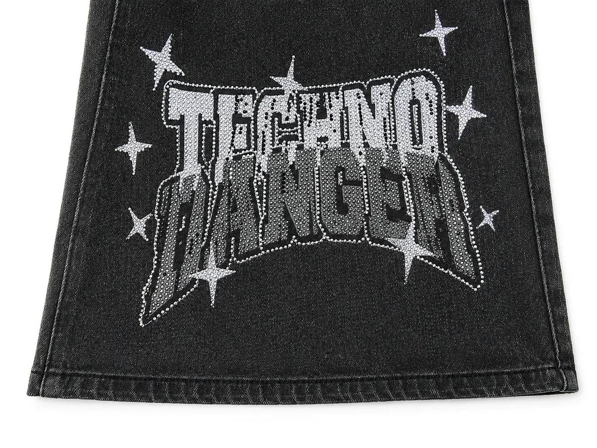 A close-up of a black denim garment, likely a skirt or shorts, featuring a large arched 'TECHNO DANGER' graphic embellished with rhinestones and surrounded by white star accents. TCH Techno Magic Star Graphic Wide-Leg Jeans