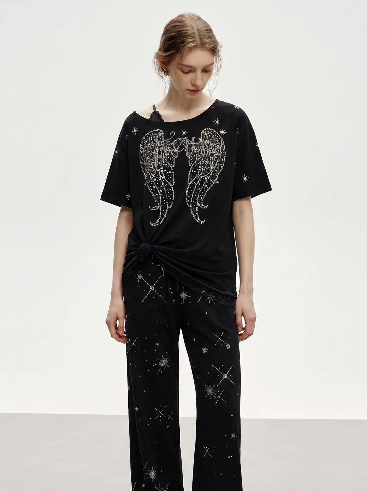 A model wearing a black two-piece lounge set featuring a short-sleeve top with a large rhinestone angel wings graphic and matching wide-leg pants with a rhinestone star pattern, set against a plain white studio background. TCH Techno Rhinestone Angel Wings Lace Trim Off-Shoulder Top