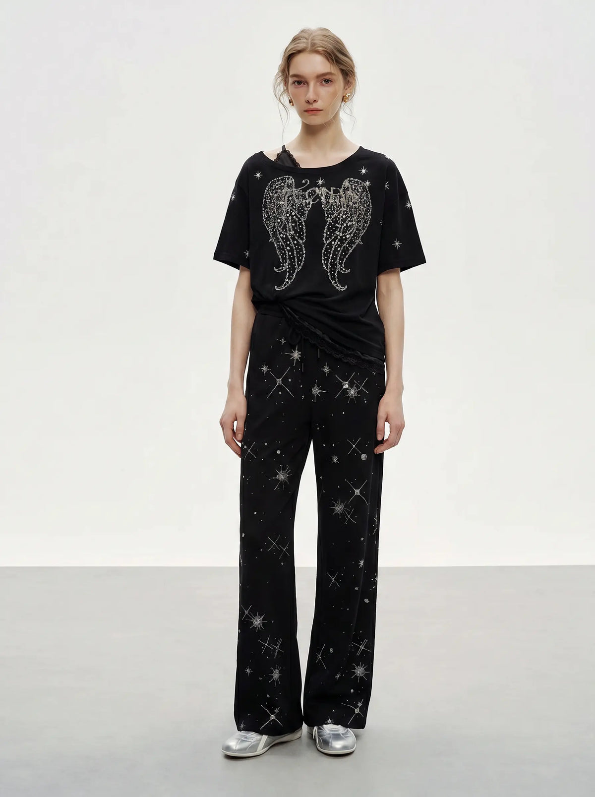 A model stands in a studio wearing a black short-sleeve top with a silver wing graphic and matching black wide-leg pants covered in small silver star prints, paired with metallic silver shoes. TCH Techno Rhinestone Angel Wings Lace Trim Off-Shoulder Top