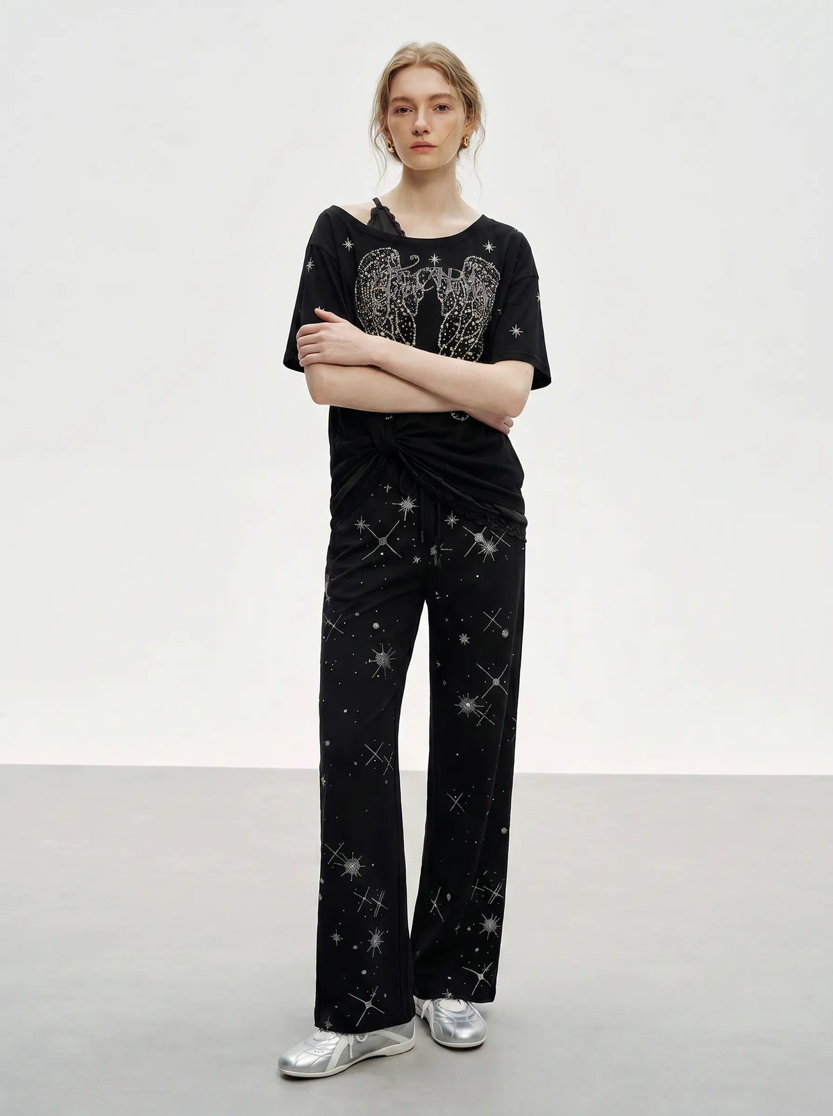 A model stands in a studio wearing a black short-sleeve top and matching wide-leg pants, both decorated with silver rhinestone star and wing patterns, paired with silver sneakers. TCH Techno Rhinestone Angel Wings Lace Trim Off-Shoulder Top