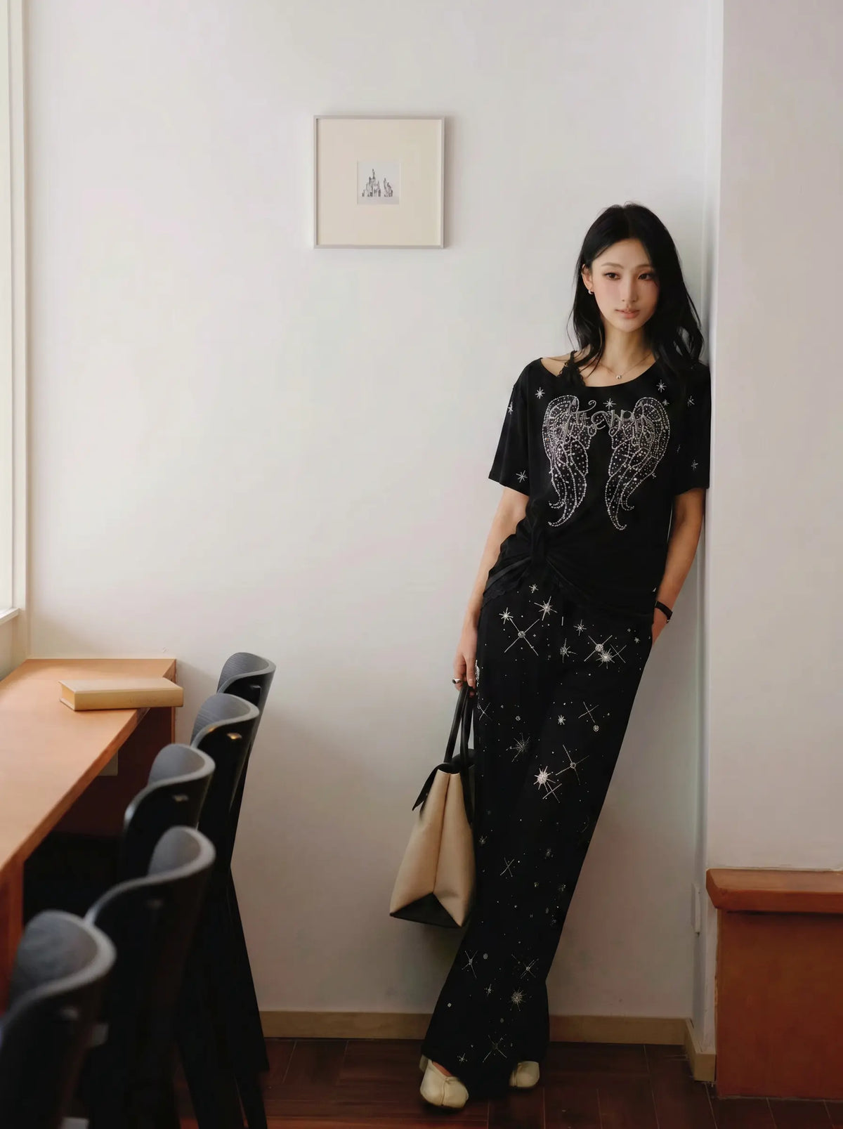 A woman stands indoors wearing a black two-piece outfit with a wing graphic on the top and star-like sparkles on the pants, holding a beige tote bag. TCH Techno Rhinestone Angel Wings Lace Trim Off-Shoulder Top