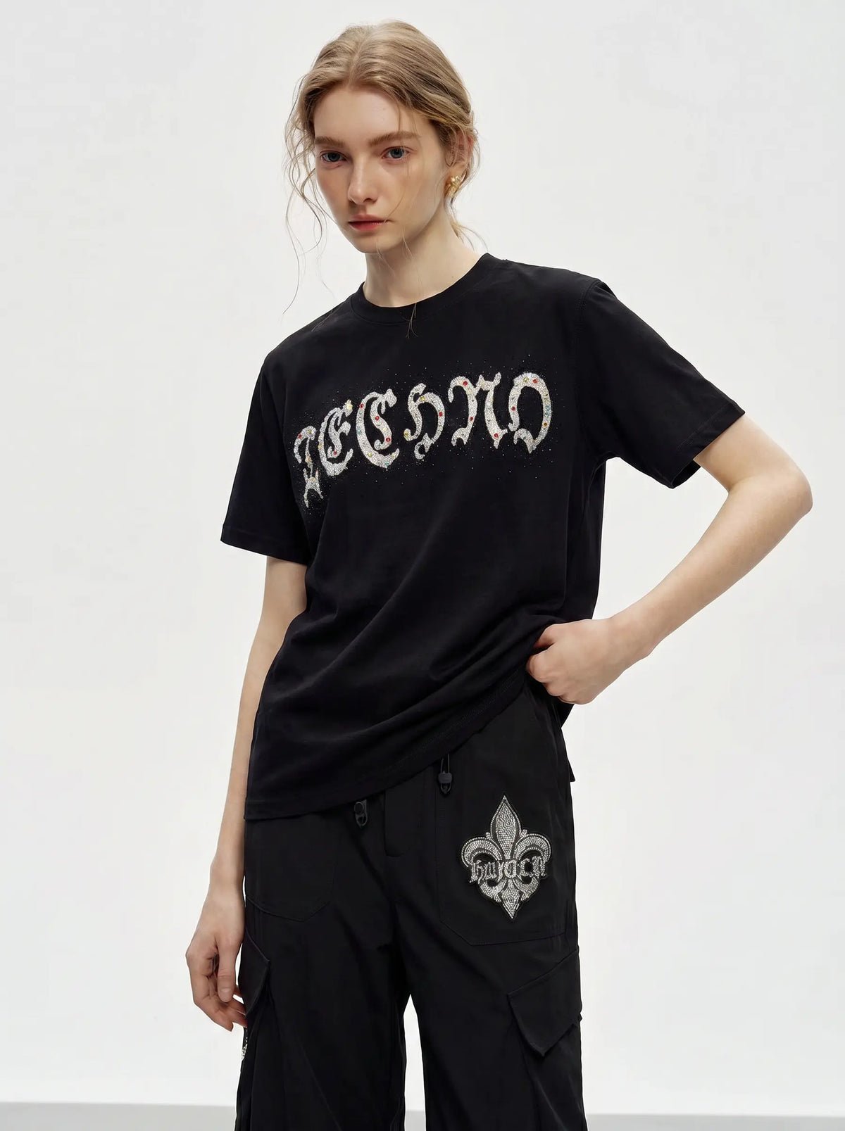 A model wearing a black short-sleeve t-shirt with a white gothic-style 'REIGN' graphic across the chest, paired with black cargo pants featuring a white fleur-de-lis graphic on the pocket, set against a plain light gray studio background. TCH Techno Rhinestone Gothic Graphic Black T-Shirt