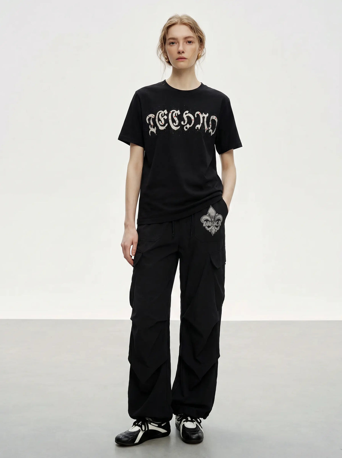 A model stands in a studio wearing a black short-sleeve T-shirt with a white gothic-style graphic across the chest, black cargo pants with a white graphic detail on the pocket, and black-and-white sneakers. TCH Techno Rhinestone Gothic Graphic Black T-Shirt