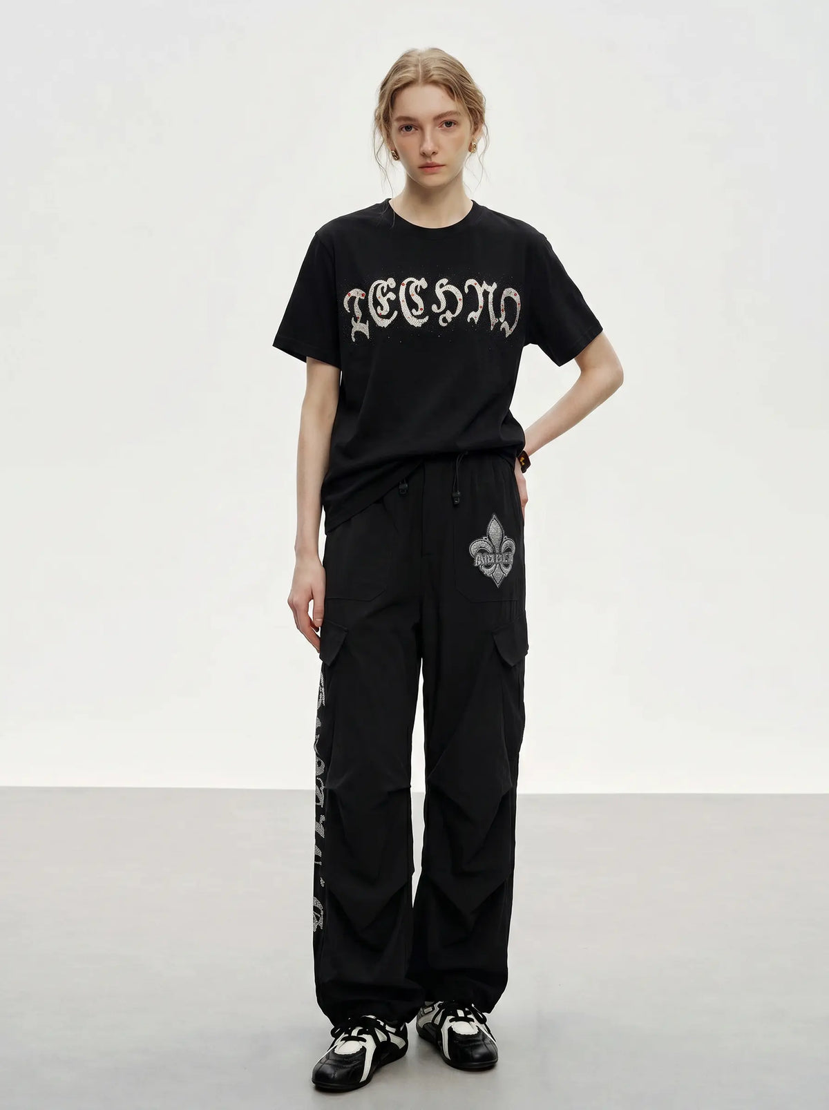 A model stands in a studio wearing a black graphic T-shirt with a gothic-style white logo and black wide-leg cargo pants with a white fleur-de-lis graphic on the thigh. TCH Techno Rhinestone Gothic Graphic Black T-Shirt