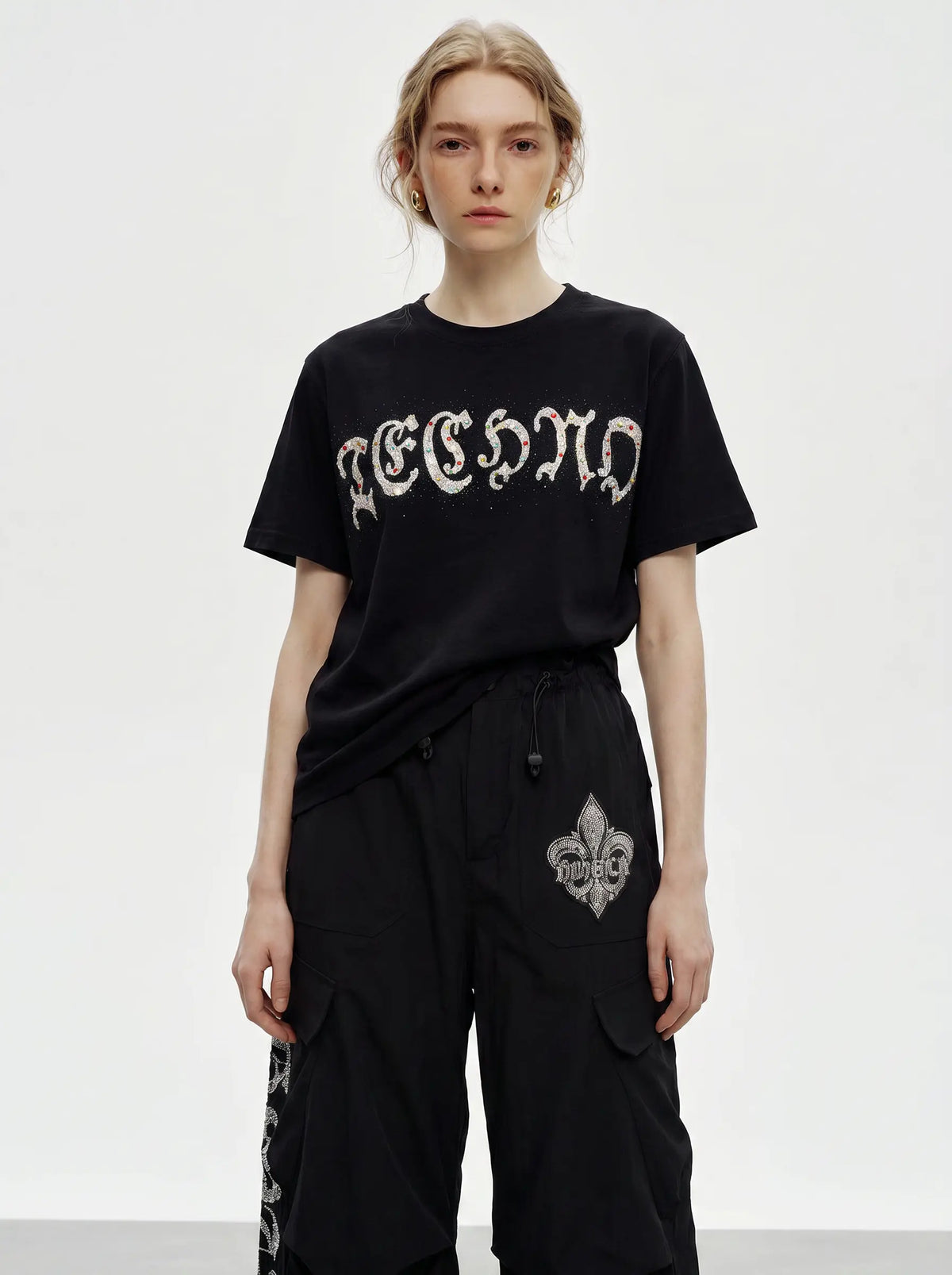 A model wearing a black short-sleeve t-shirt with a distressed white gothic-style graphic reading 'RECYCLED' across the chest, paired with black cargo pants featuring a white fleur-de-lis skull graphic on the thigh. TCH Techno Rhinestone Gothic Graphic Black T-Shirt