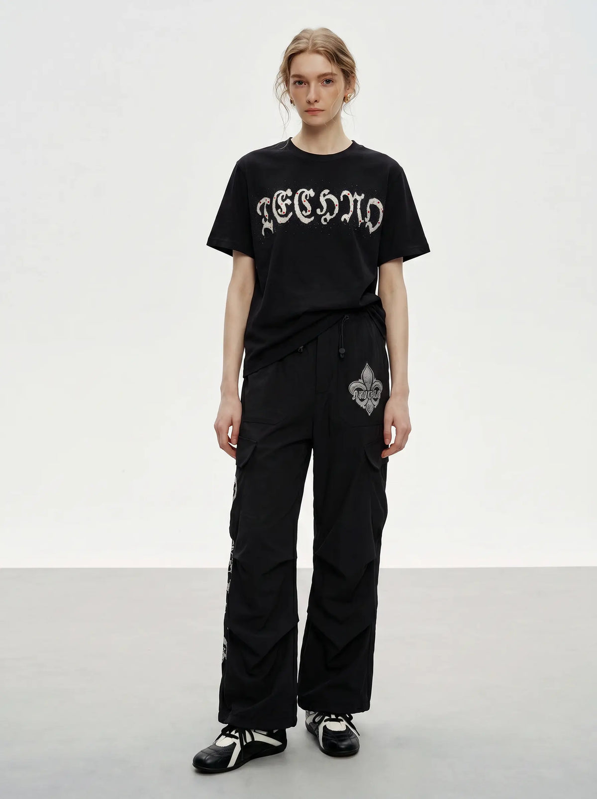 A model stands in a studio wearing a black t-shirt with a white gothic-style graphic print and black wide-leg cargo pants with white graphic details. TCH Techno Rhinestone Gothic Graphic Black T-Shirt