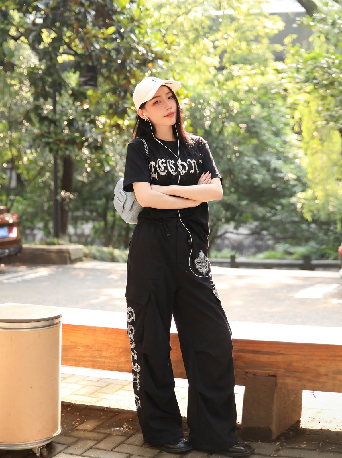 A woman stands outdoors on a bench in a park-like setting, wearing a black graphic T-shirt, black wide-leg cargo pants with white graphic prints down the sides, a white baseball cap, and white earphones. TCH Techno Rhinestone Gothic Graphic Black T-Shirt