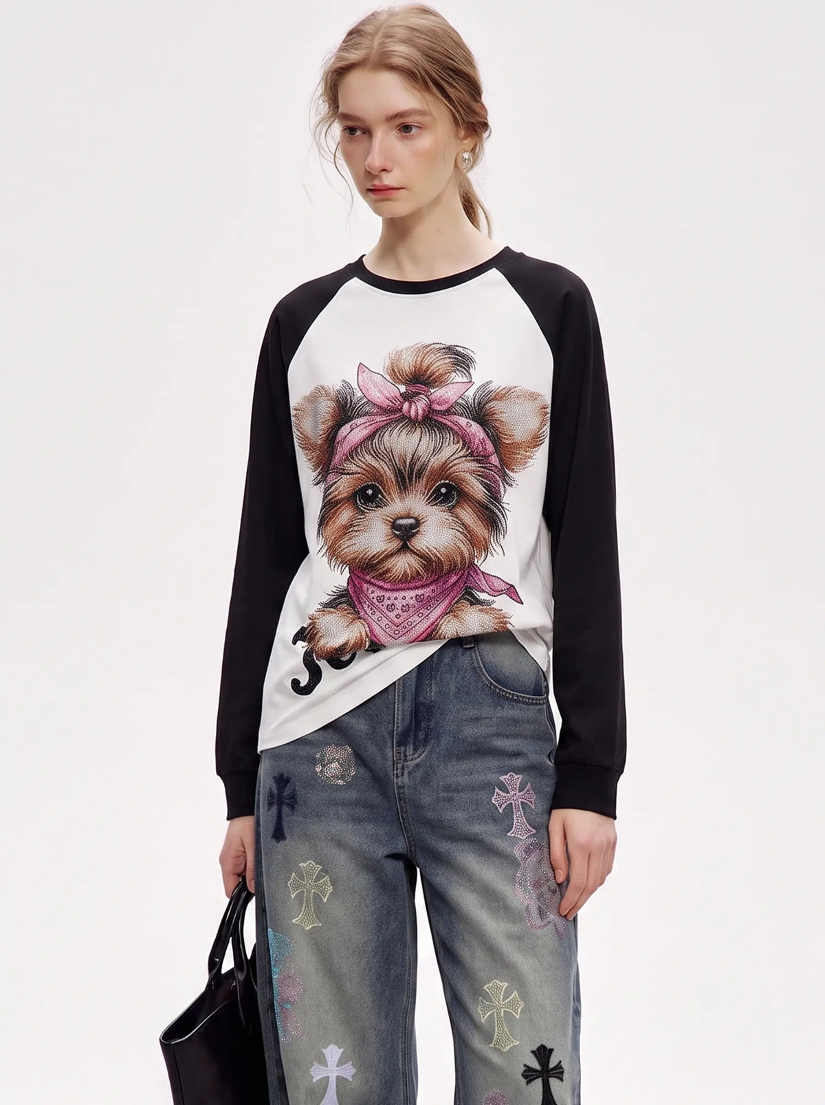 A model wearing a white and black raglan long-sleeve shirt with a detailed graphic of a Yorkshire Terrier wearing a pink bandana and bow. She is paired with distressed blue jeans featuring cross patches and is holding a black handbag against a plain white studio background. TCH Techno Teddy Graphic Raglan Long Sleeve Shirt