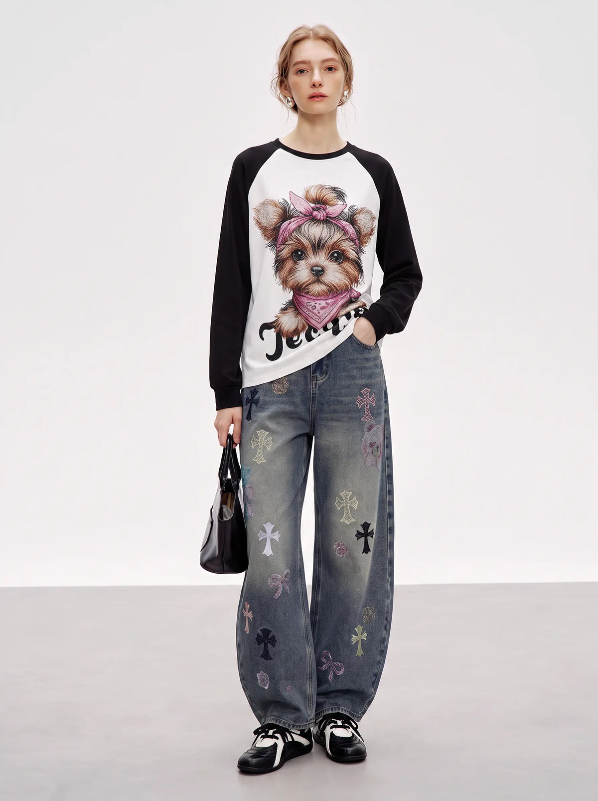 A model stands against a light gray studio background wearing a black-and-white raglan long-sleeve top with a large dog graphic on the front, loose gray jeans with embroidered cross motifs, black-and-white sneakers, and holding a small black handbag. TCH Techno Teddy Graphic Raglan Long Sleeve Shirt