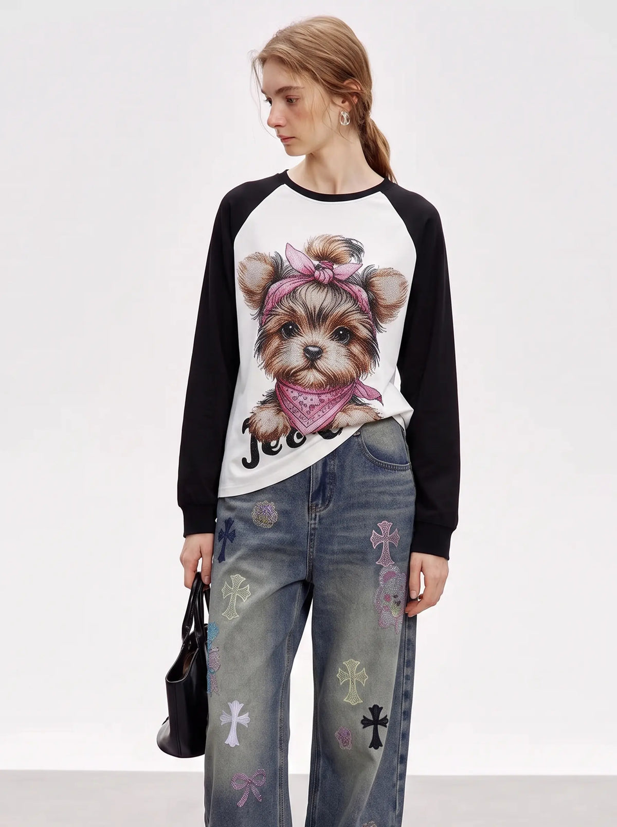 A model wearing a black and white raglan long-sleeve shirt with a detailed graphic of a Yorkshire Terrier wearing a pink bandana, paired with distressed blue jeans featuring cross and bow embroidery, holding a small black handbag against a plain white studio background. TCH Techno Teddy Graphic Raglan Long Sleeve Shirt