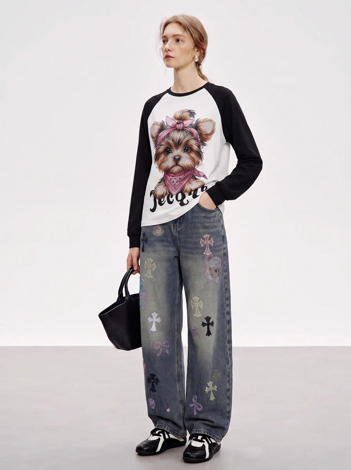 A model stands in a studio wearing a black and white raglan long-sleeve shirt with a graphic of a dog wearing a pink bow and bandana, paired with wide-leg gray jeans decorated with small cross motifs, black sneakers, and holding a black handbag. TCH Techno Teddy Graphic Raglan Long Sleeve Shirt