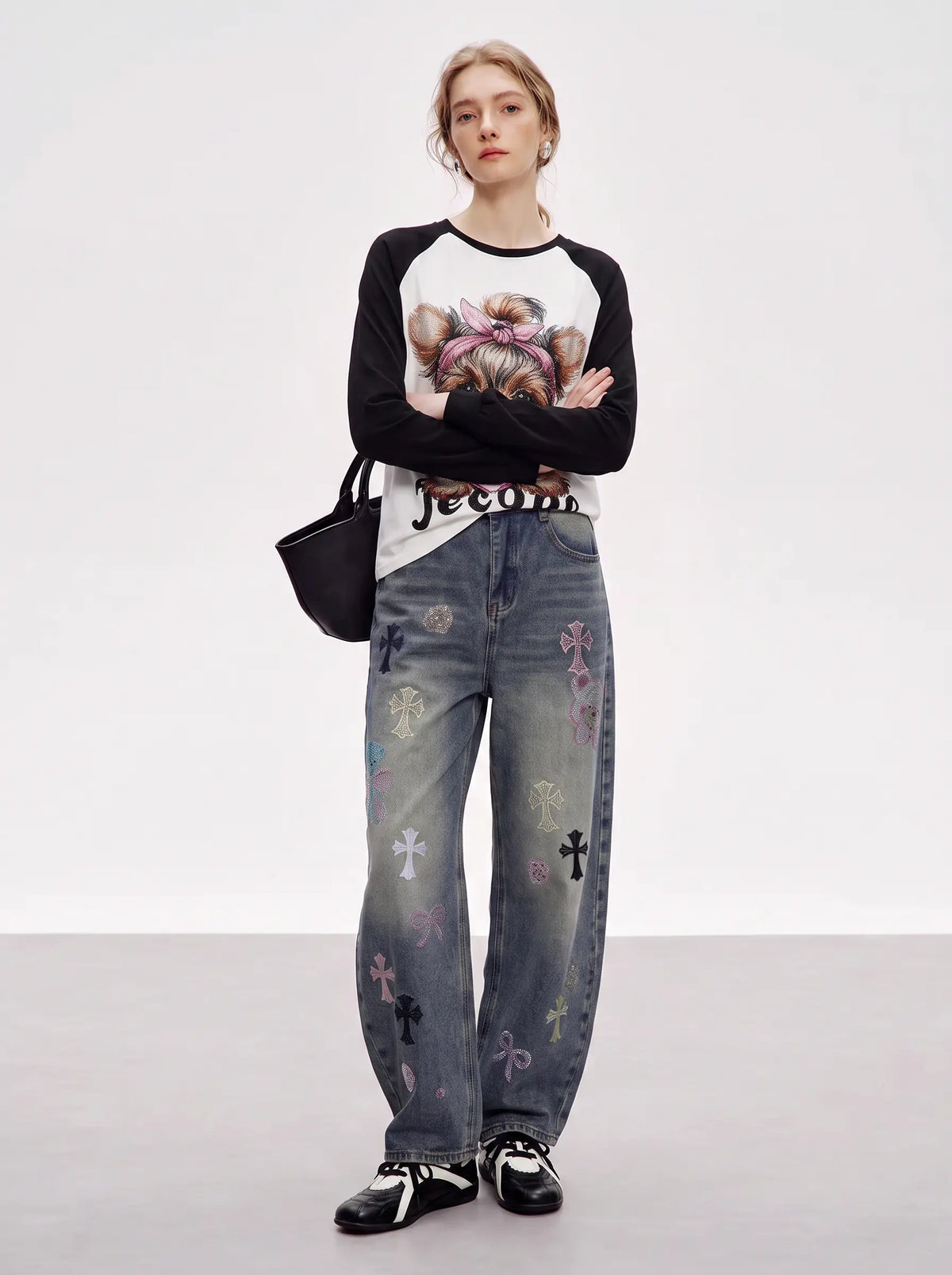 A model stands against a light gray studio background wearing a black and white long-sleeve graphic top with a teddy bear print, wide-leg gray jeans with colorful embroidered crosses, a black belt, and black-and-white sneakers. She is holding a black shoulder bag. TCH Techno Teddy Graphic Raglan Long Sleeve Shirt