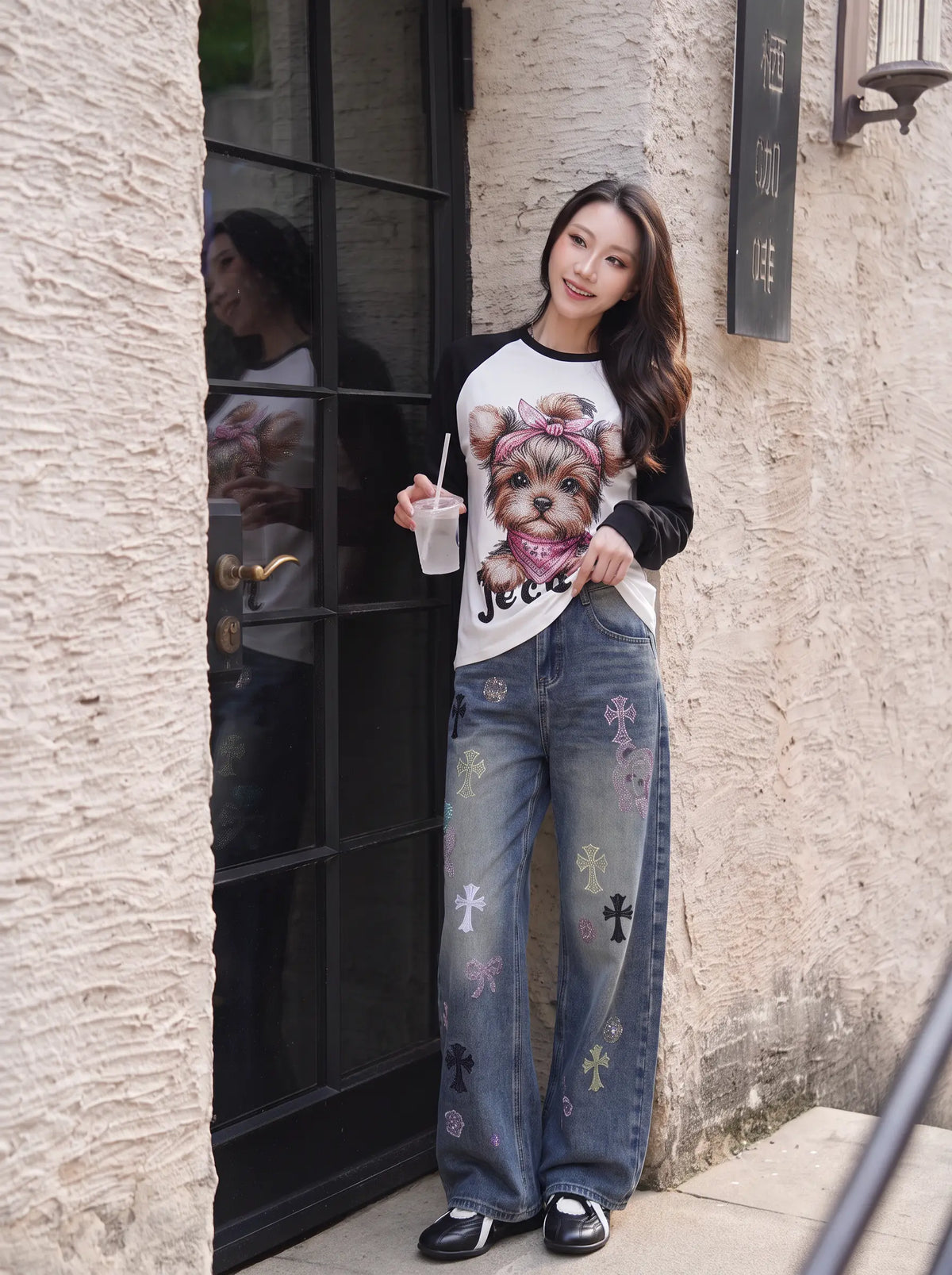 A woman stands outdoors in front of a textured beige wall and black-framed glass door, wearing a white graphic T-shirt with a dog illustration and the word 'Jeep' on the front, paired with wide-leg blue jeans featuring colorful cross motifs and black loafers. TCH Techno Teddy Graphic Raglan Long Sleeve Shirt