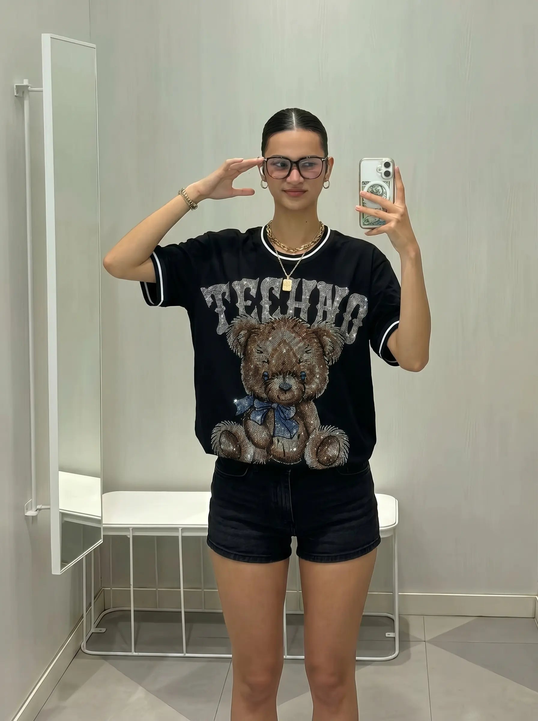 Person wearing a black t-shirt with a teddy bear graphic and text, standing in a bathroom. TCH Techno Teddy Rhinestone T-Shirt