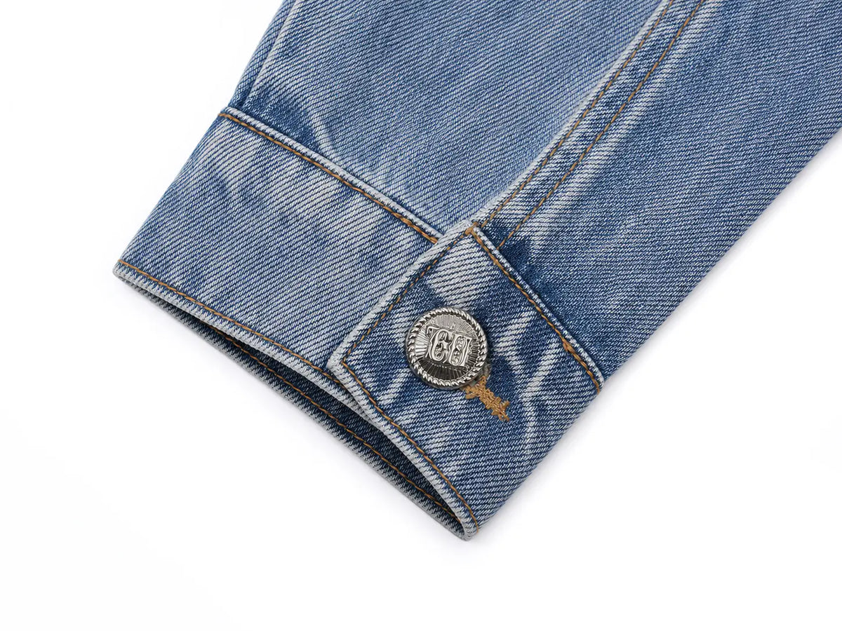 Close-up of a blue denim jacket cuff showing a silver metal button with a circular logo and contrast stitching on a white background. TCH Techno Wings Rhinestone Graphic Denim Jacket