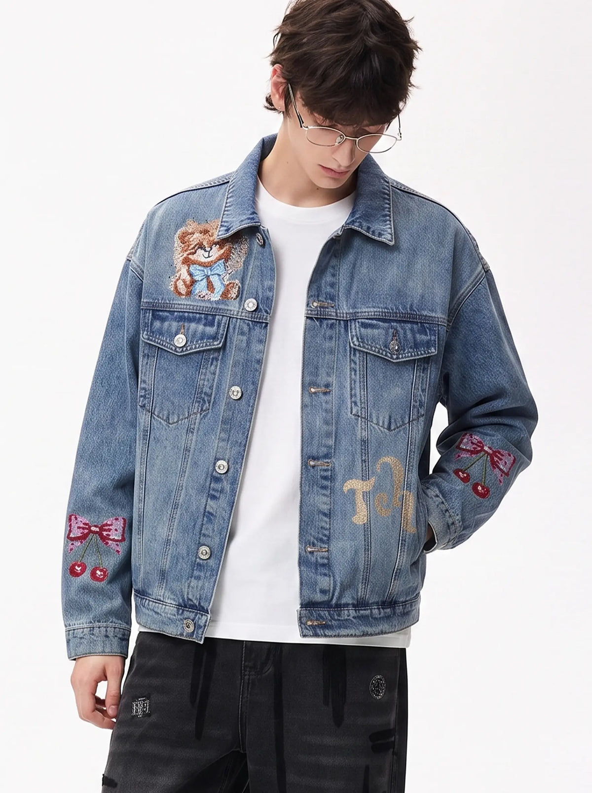 A model wearing a light blue denim jacket with various embroidered patches, including a teddy bear on the left chest and cherries on the sleeve, styled over a white t-shirt and dark pants. TCH Techno Wings Rhinestone Graphic Denim Jacket