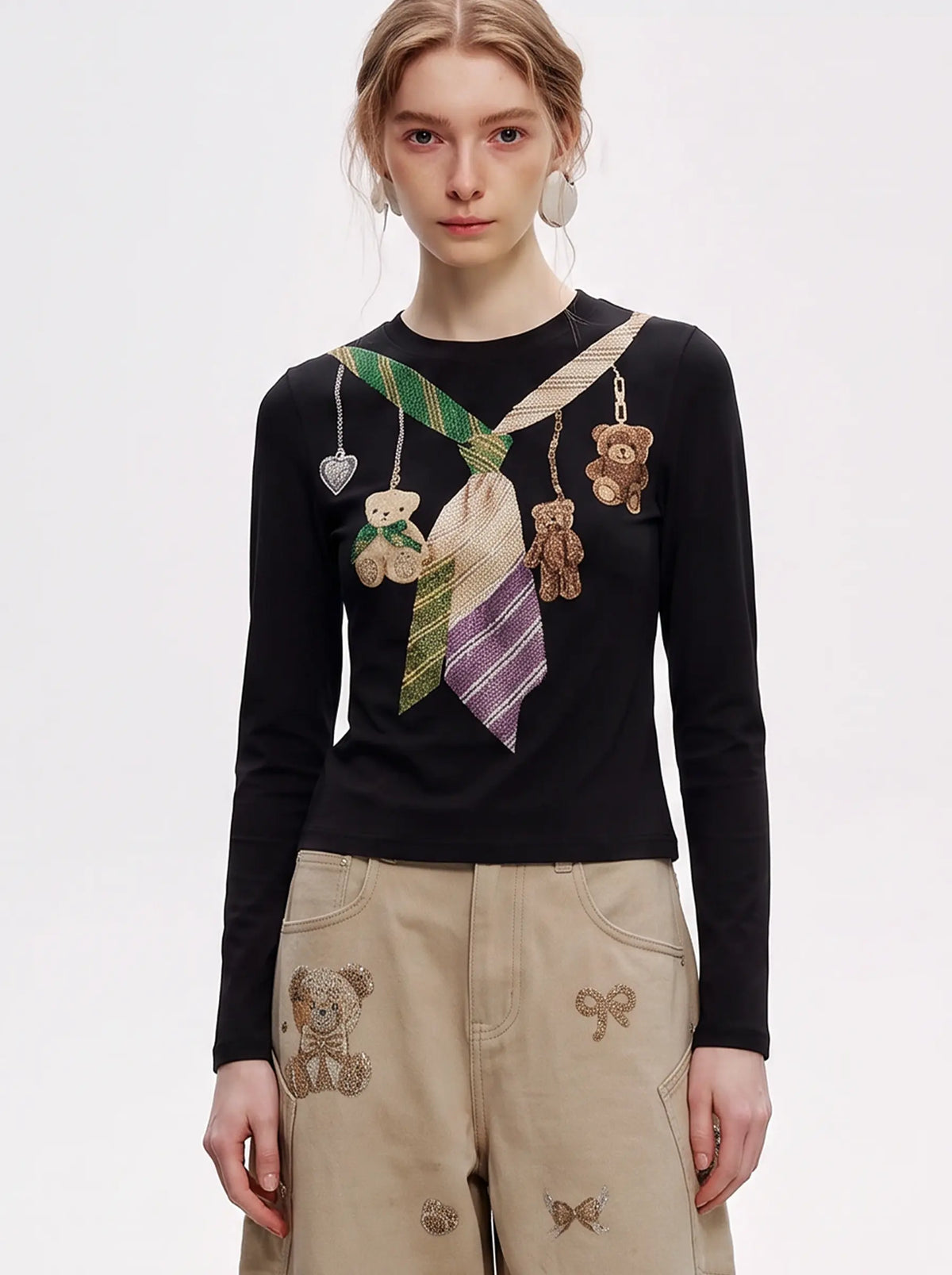 A model wearing a black long-sleeve top with a colorful necktie and teddy bear graphic print, paired with beige cargo pants featuring teddy bear embroidery, against a plain white studio background. TCH Teddy Bear Charm Tie Graphic Black Long Sleeve T-Shirt