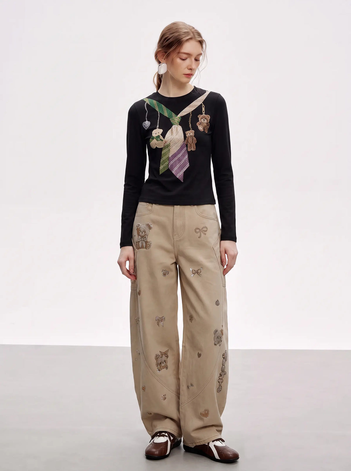 A model stands in a studio wearing a black long-sleeve top with a colorful tie and teddy bear graphic, paired with beige wide-leg trousers featuring teddy bear prints, and brown shoes. TCH Teddy Bear Charm Tie Graphic Black Long Sleeve T-Shirt