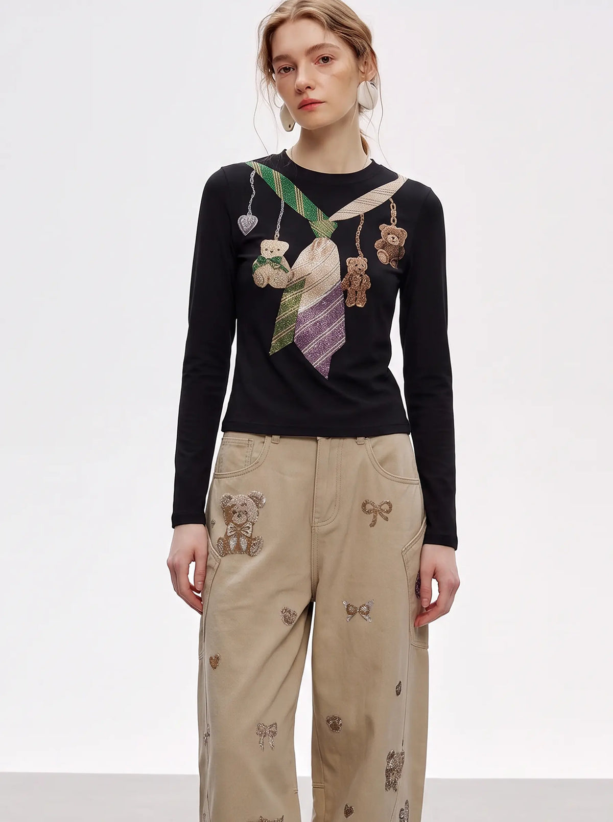 A model wearing a black long-sleeve crewneck top with a colorful embroidered tie and teddy bear motif on the chest, paired with beige wide-leg trousers featuring teddy bear and bow embroidery. TCH Teddy Bear Charm Tie Graphic Black Long Sleeve T-Shirt