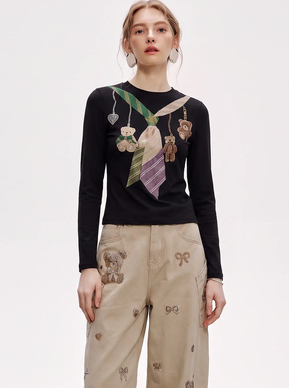A model wearing a black long-sleeve knit top with a colorful embroidered necktie and teddy bear motif, paired with beige wide-leg pants featuring teddy bear and bow embroidery, against a plain white studio background. TCH Teddy Bear Charm Tie Graphic Black Long Sleeve T-Shirt