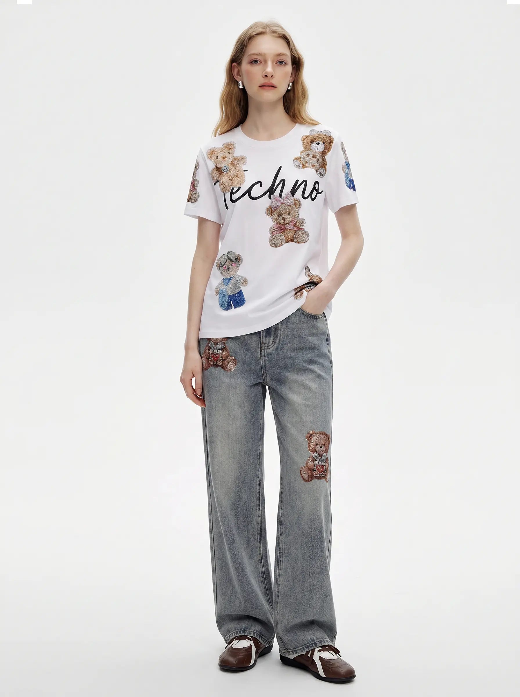 A model wearing a white short-sleeve t-shirt with teddy bear patches and black script text, paired with light gray wide-leg jeans featuring teddy bear patches on the thighs and knees, and brown loafers, against a plain white studio background. TCH Teddy Bear Rhinestone All-Over Graphic T-Shirt
