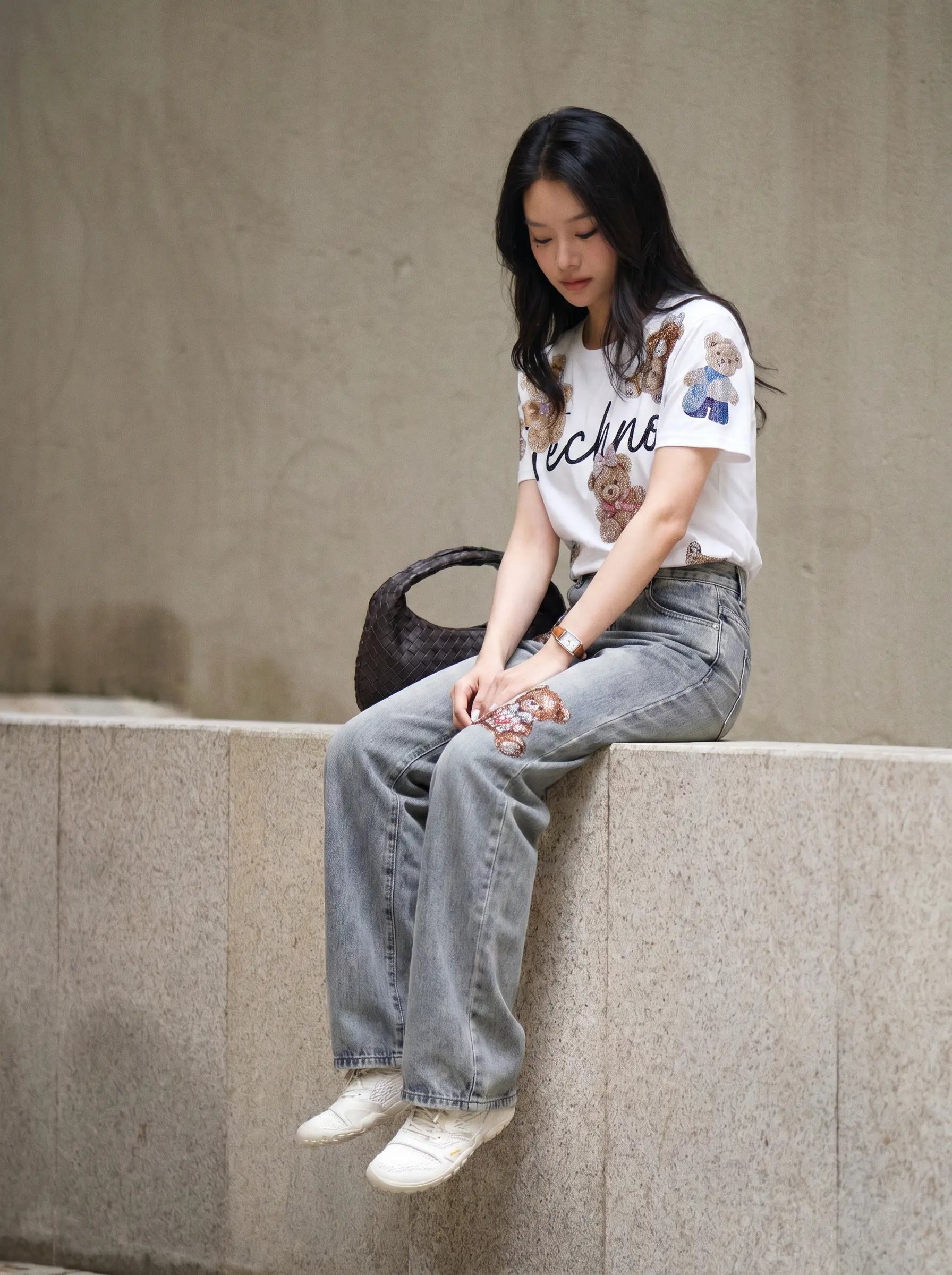 A woman sits on a concrete ledge wearing a white graphic t-shirt with teddy bear prints and the word 'techno', paired with light-wash wide-leg jeans and white sneakers. TCH Teddy Bear Rhinestone All-Over Graphic T-Shirt