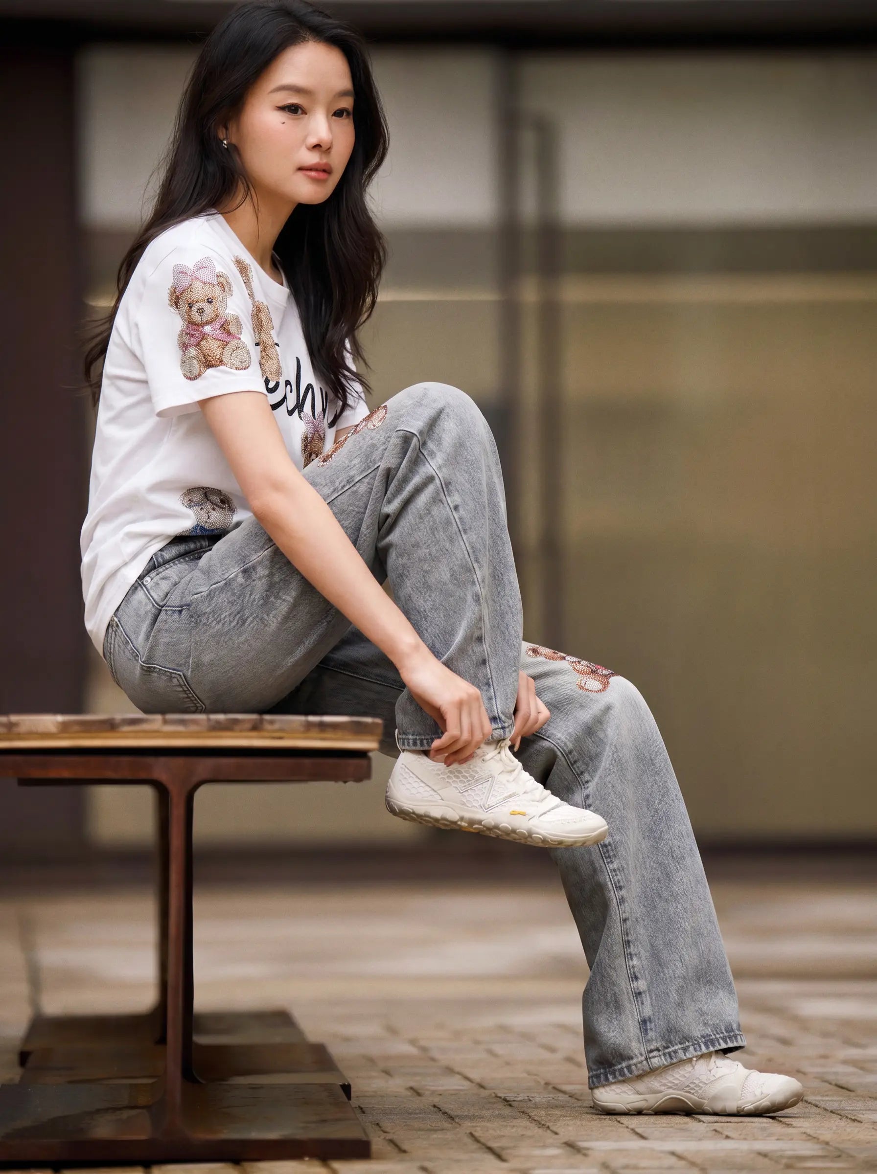 A woman sits on a wooden bench wearing a white graphic t-shirt with a teddy bear print, light-wash distressed jeans, and off-white sneakers in an outdoor urban setting. TCH Teddy Bear Rhinestone All-Over Graphic T-Shirt