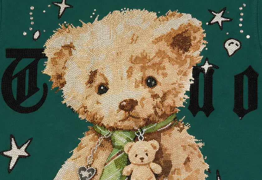 A close-up of a teddy bear cross-stitch or needlepoint design on a dark teal fabric background, featuring a large beige teddy bear with a green bow tie and a smaller teddy bear charm, with black decorative letters and white stars around it. TCH Teddy Bear Rhinestone Graphic T-Shirt