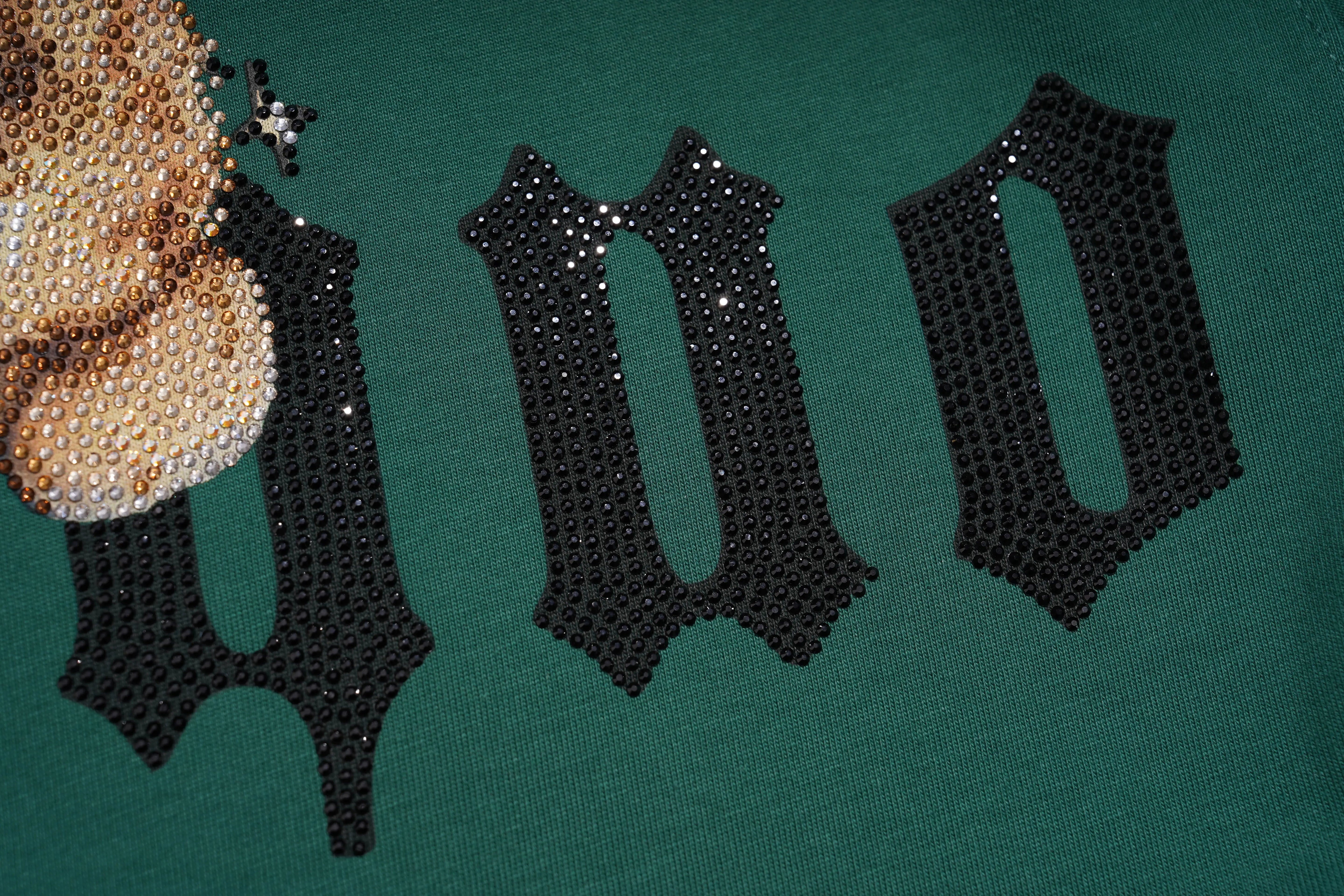 Close-up of a green sweatshirt featuring large black Gothic-style letters embellished with rhinestones, alongside a gold rhinestone graphic on the left. TCH Teddy Bear Rhinestone Graphic T-Shirt