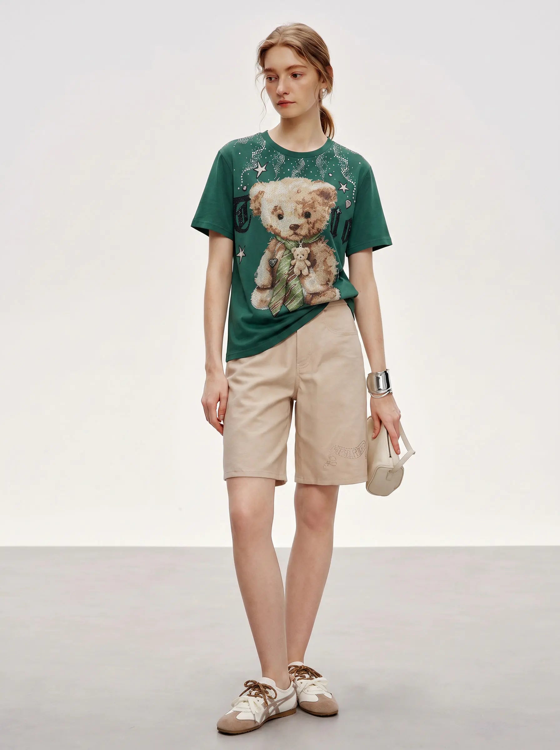 A woman modeling a dark green graphic t-shirt with a teddy bear print, paired with beige shorts and white sneakers, standing against a neutral studio background. TCH Teddy Bear Rhinestone Graphic T-Shirt