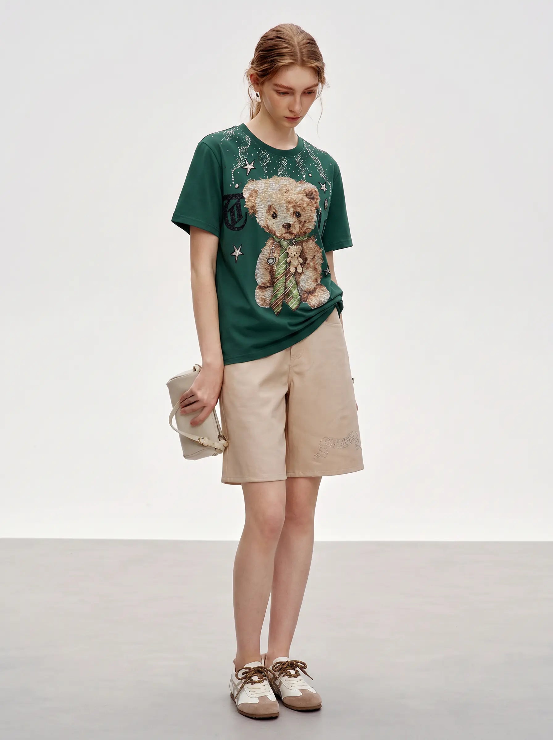 A model stands in a studio wearing a dark green graphic T-shirt with a teddy bear print, beige cargo shorts, and brown-and-white shoes, while holding a small beige clutch. TCH Teddy Bear Rhinestone Graphic T-Shirt