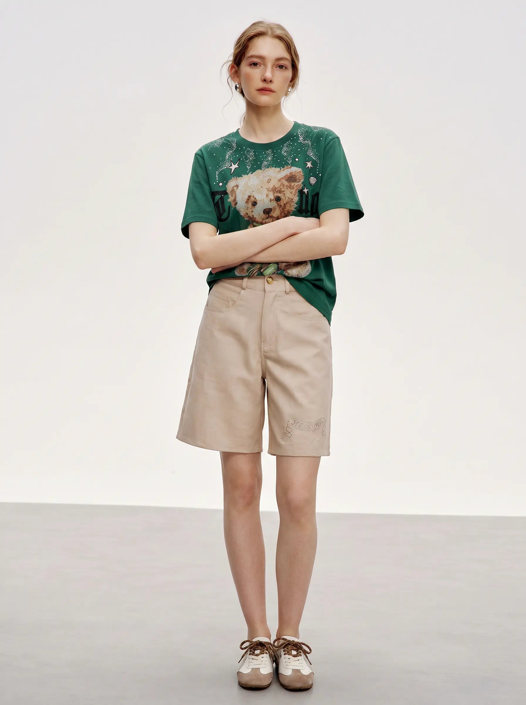 A model stands in a studio wearing a green graphic T-shirt with a teddy bear print, beige shorts, and brown-and-white sneakers against a plain light gray background. TCH Teddy Bear Rhinestone Graphic T-Shirt