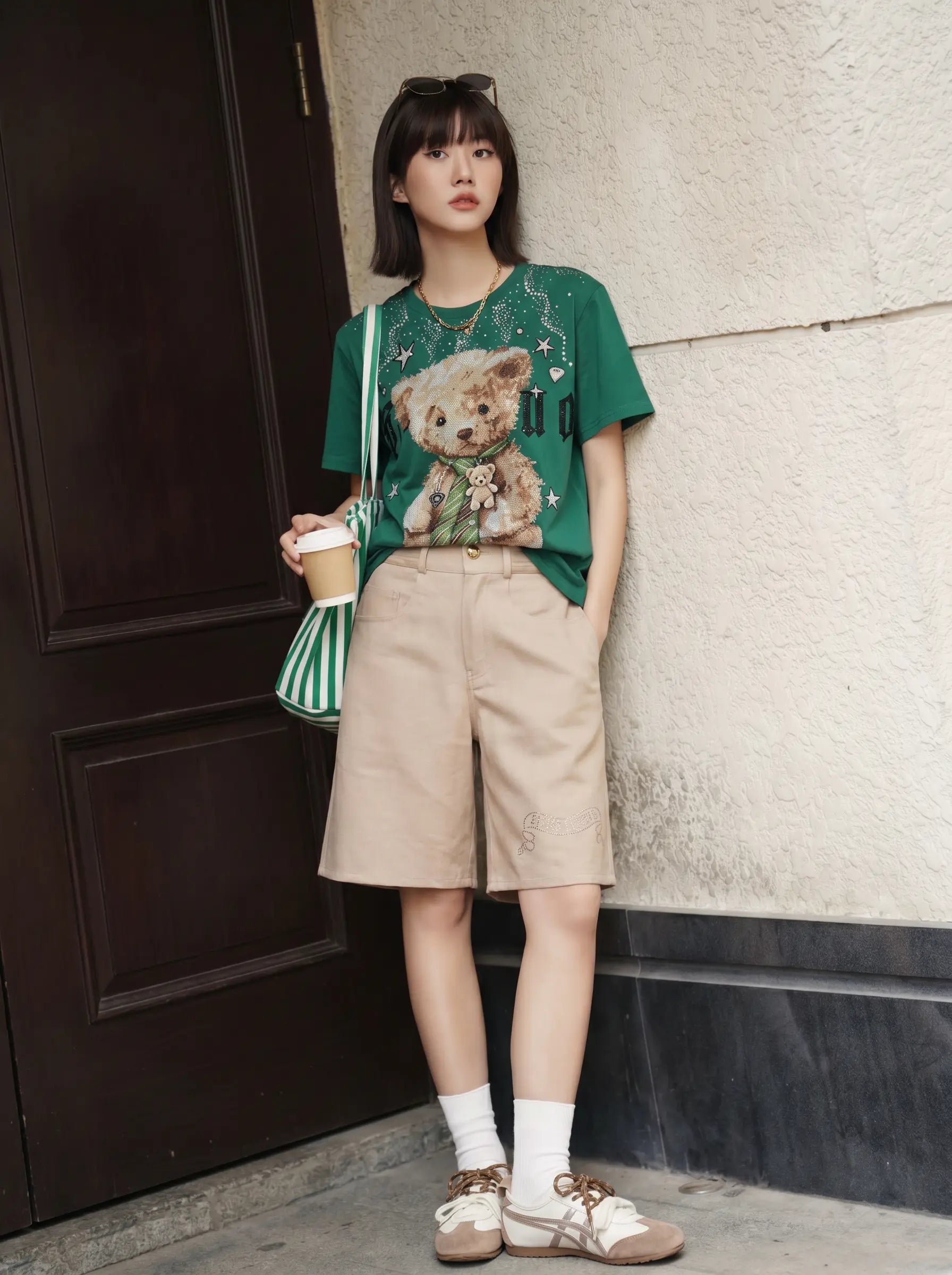 A woman stands outdoors against a textured wall and dark wooden door, wearing a green graphic T-shirt with a teddy bear print, beige shorts, white socks, and brown lace-up shoes, while holding a striped green and white shoulder bag and a coffee cup. TCH Teddy Bear Rhinestone Graphic T-Shirt