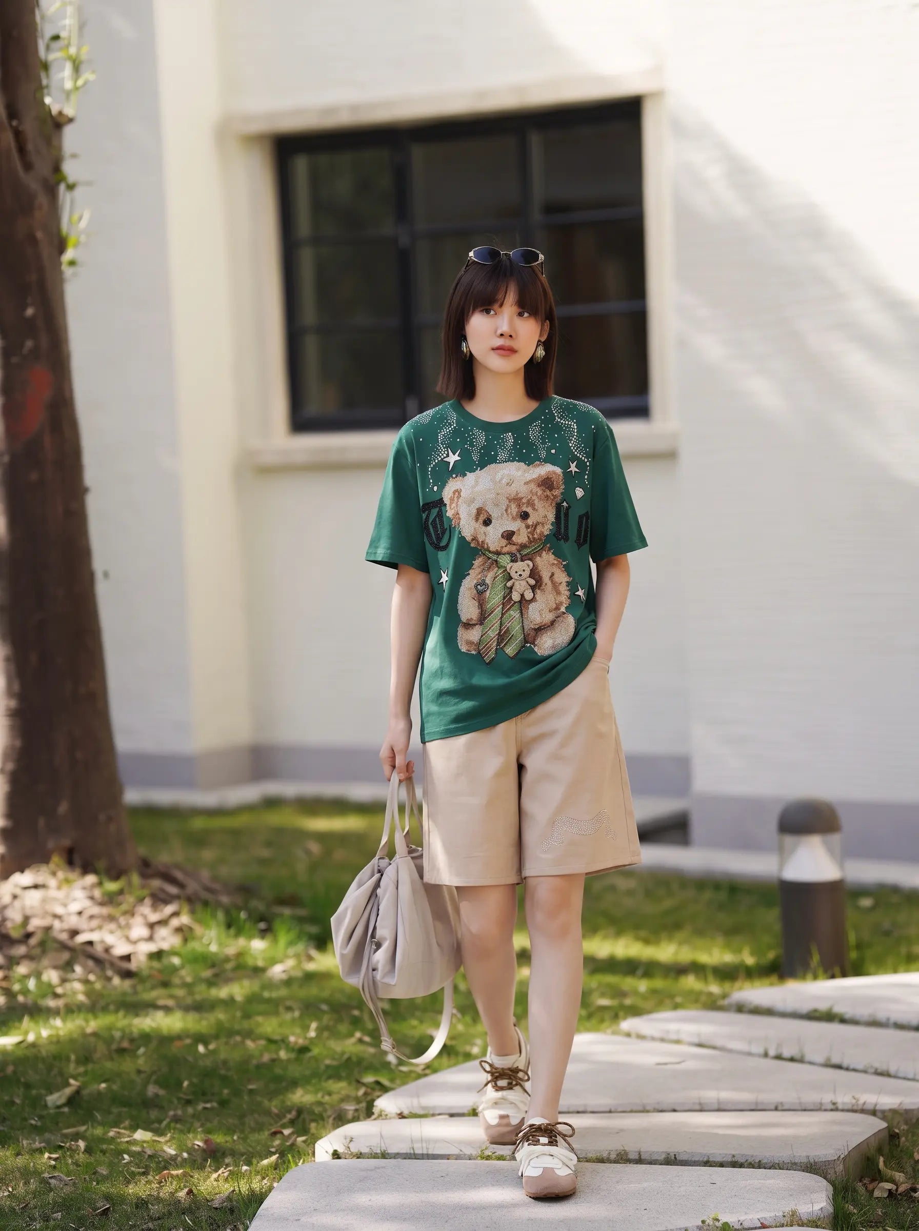 A woman stands outdoors on a stone path wearing a green graphic T-shirt with a teddy bear print, beige shorts, and carrying a light gray shoulder bag. TCH Teddy Bear Rhinestone Graphic T-Shirt