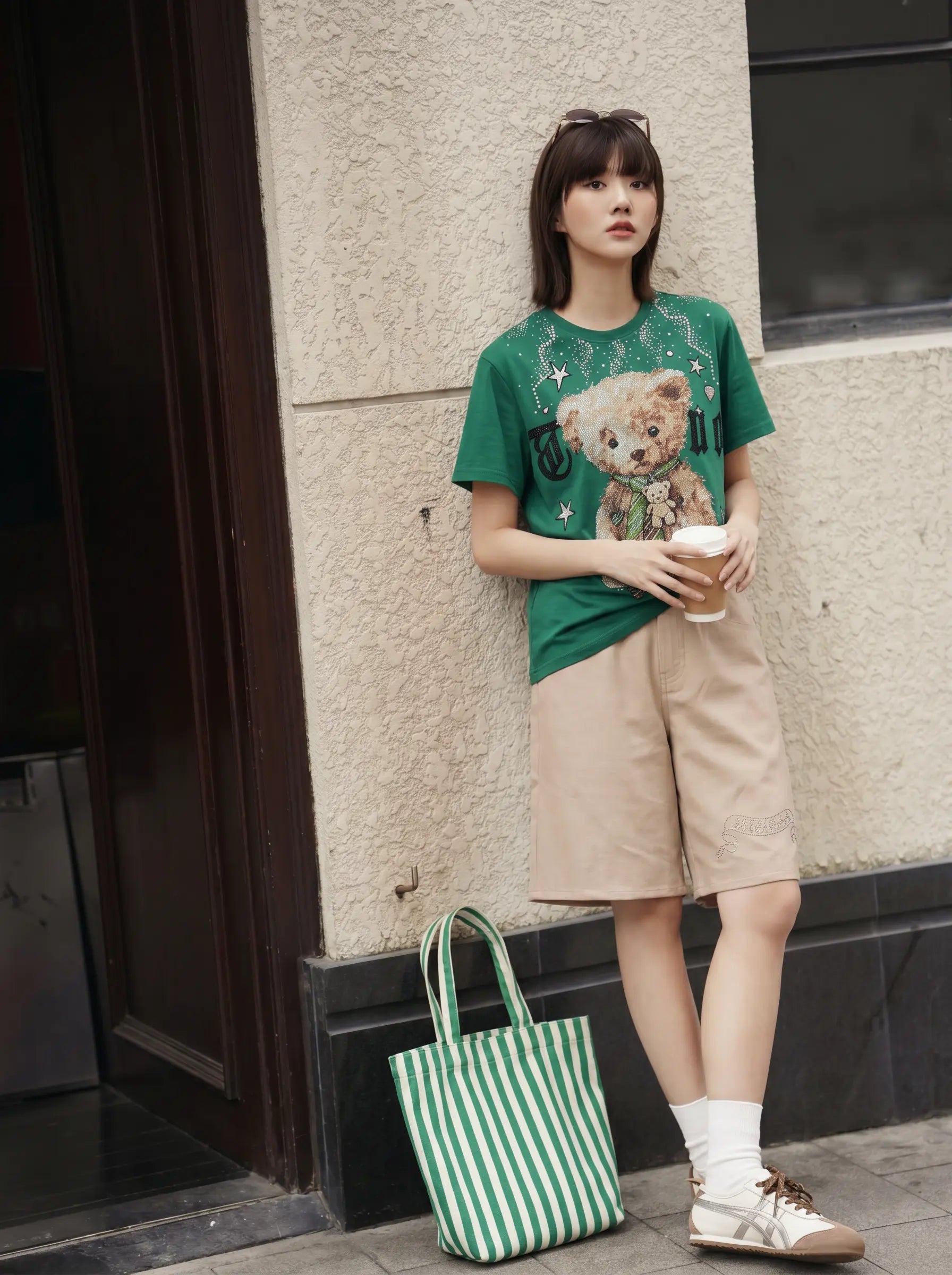 A young woman stands against a textured beige wall wearing a green graphic t-shirt with a teddy bear print, beige shorts, white socks, and sneakers. She is holding a paper coffee cup next to a green and white striped tote bag. TCH Teddy Bear Rhinestone Graphic T-Shirt