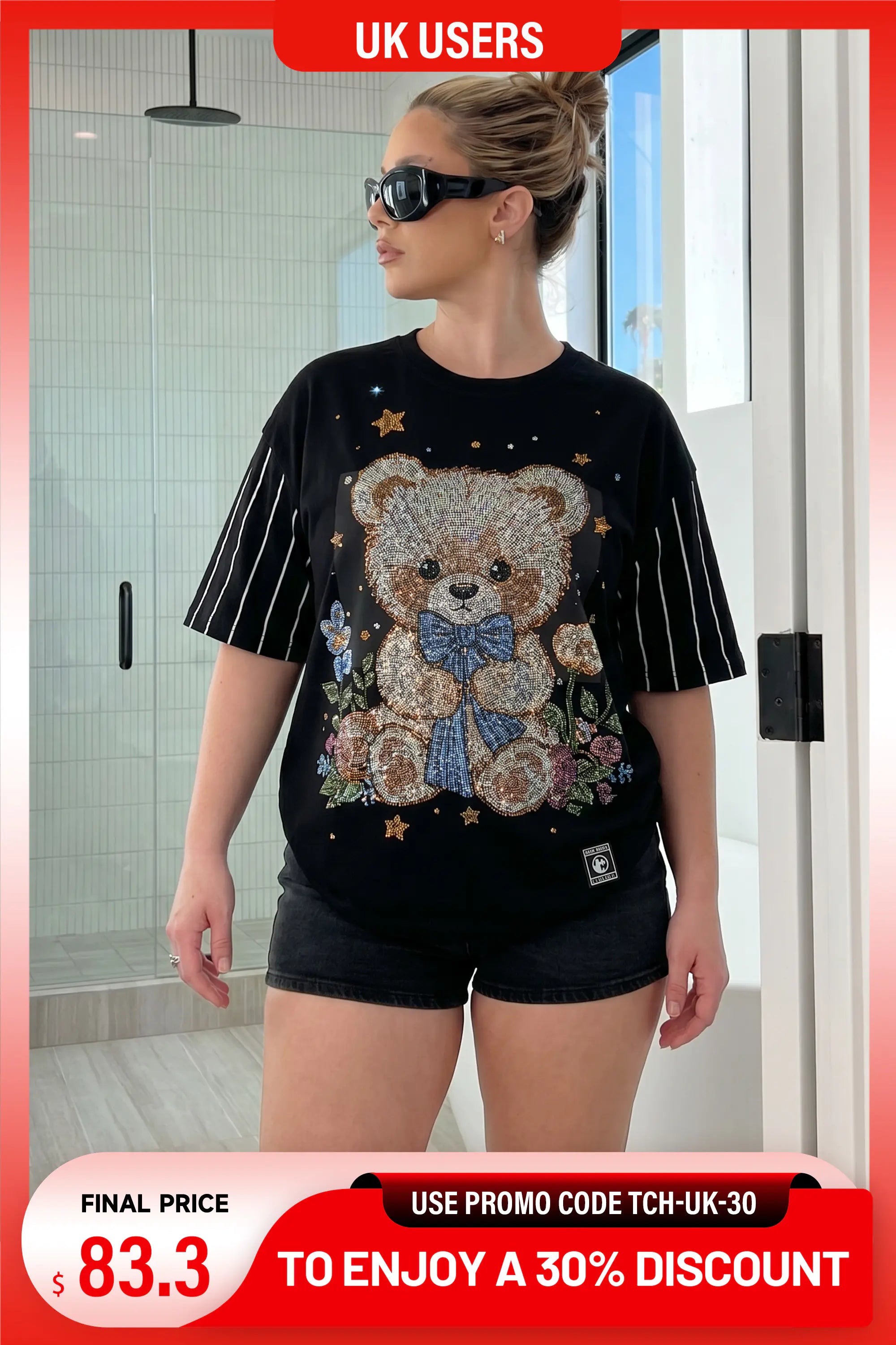 A woman wearing a black short-sleeve t-shirt with a large teddy bear graphic on the front, styled with black shorts. The t-shirt features white pinstripes on the sleeves and a teddy bear design embellished with rhinestones, set against a bathroom background. TCH Teddy Bear & Stars Rhinestone T-Shirt