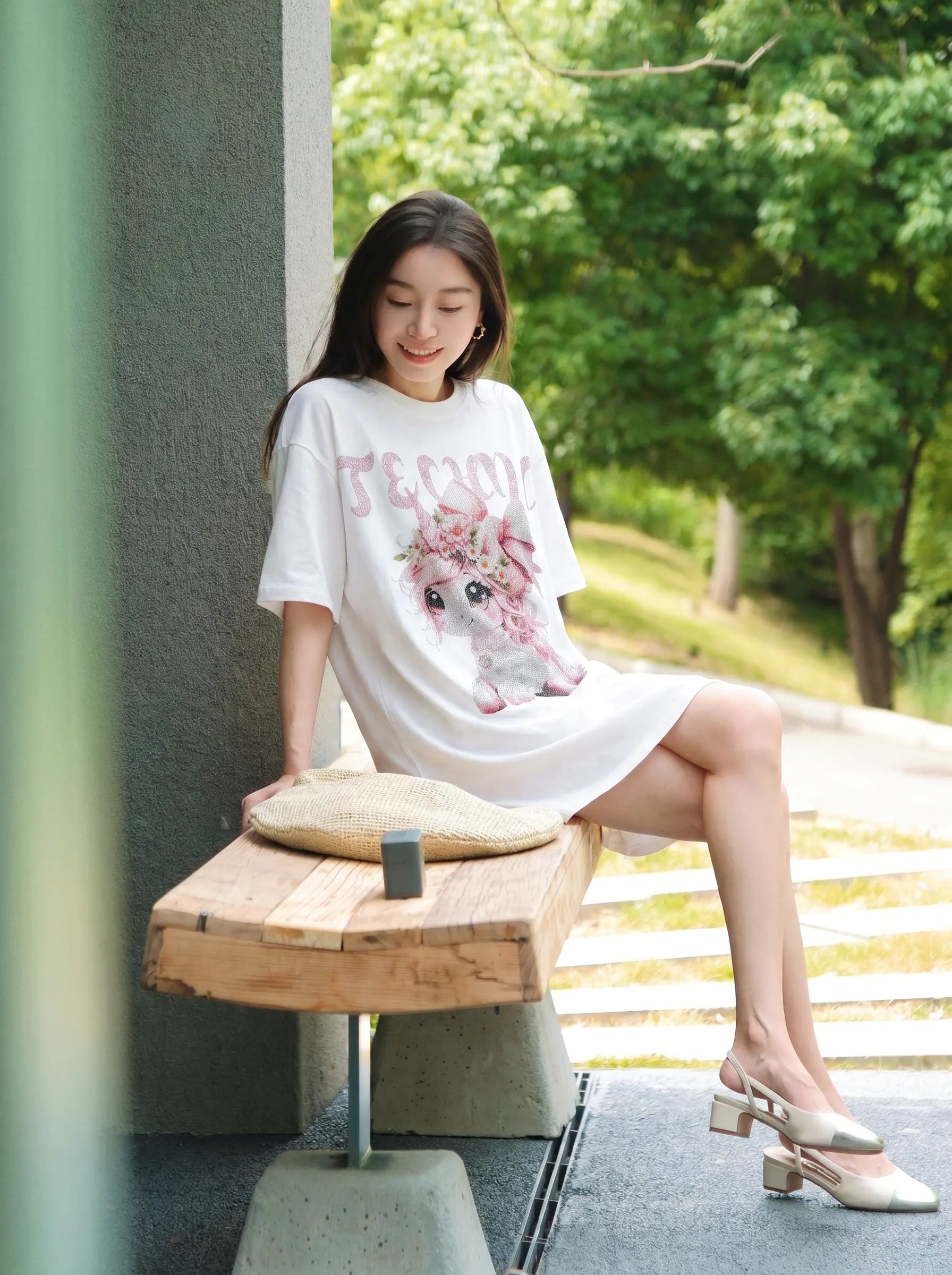 A woman sits on a wooden bench outdoors wearing a white oversized graphic T-shirt dress with a pink floral print and the word 'FEMEN' in pink. She is wearing beige block-heel mule sandals, with a woven straw hat resting on the bench in front of her. TCH Unicorn Rhinestone Angel Wings Oversized White T-Shirt
