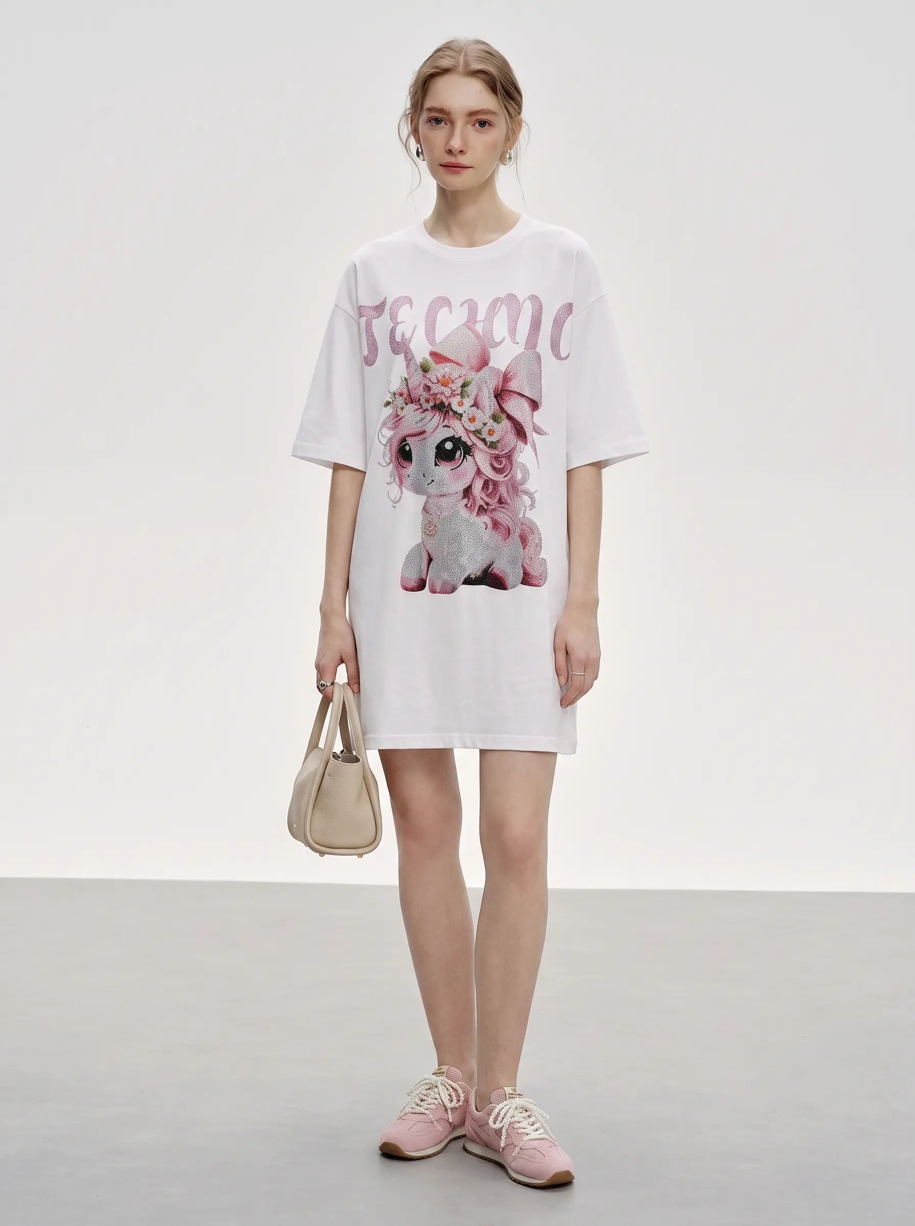 A model stands in a studio wearing a white oversized T-shirt dress with a large pink and white graphic of a flower-adorned dog and the word 'become' across the chest. She is holding a small beige handbag and wearing pink sneakers. TCH Unicorn Rhinestone Angel Wings Oversized White T-Shirt