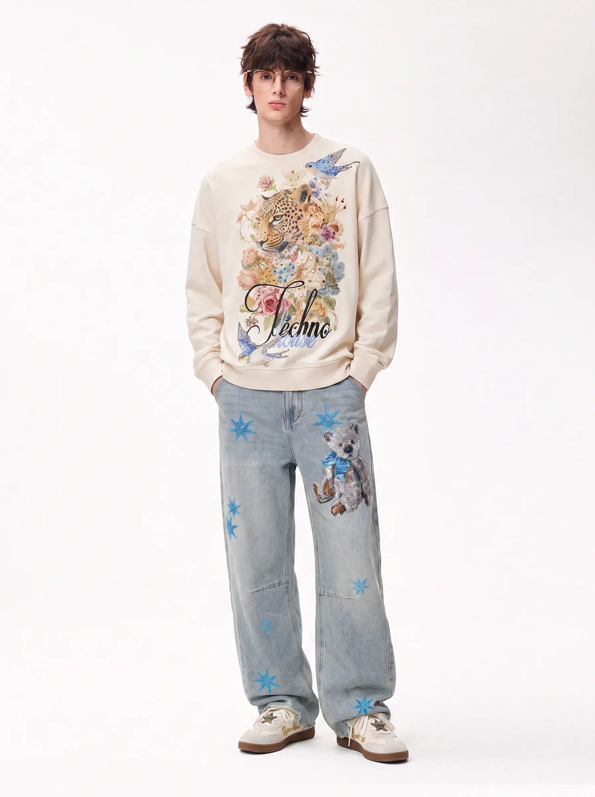 A model stands against a plain white studio background wearing a cream-colored graphic sweatshirt with a colorful floral and animal print, light-wash blue jeans with blue star patches and a bear patch, and white sneakers. TCH Vintage Leopard & Bird Graphic Sweatshirt