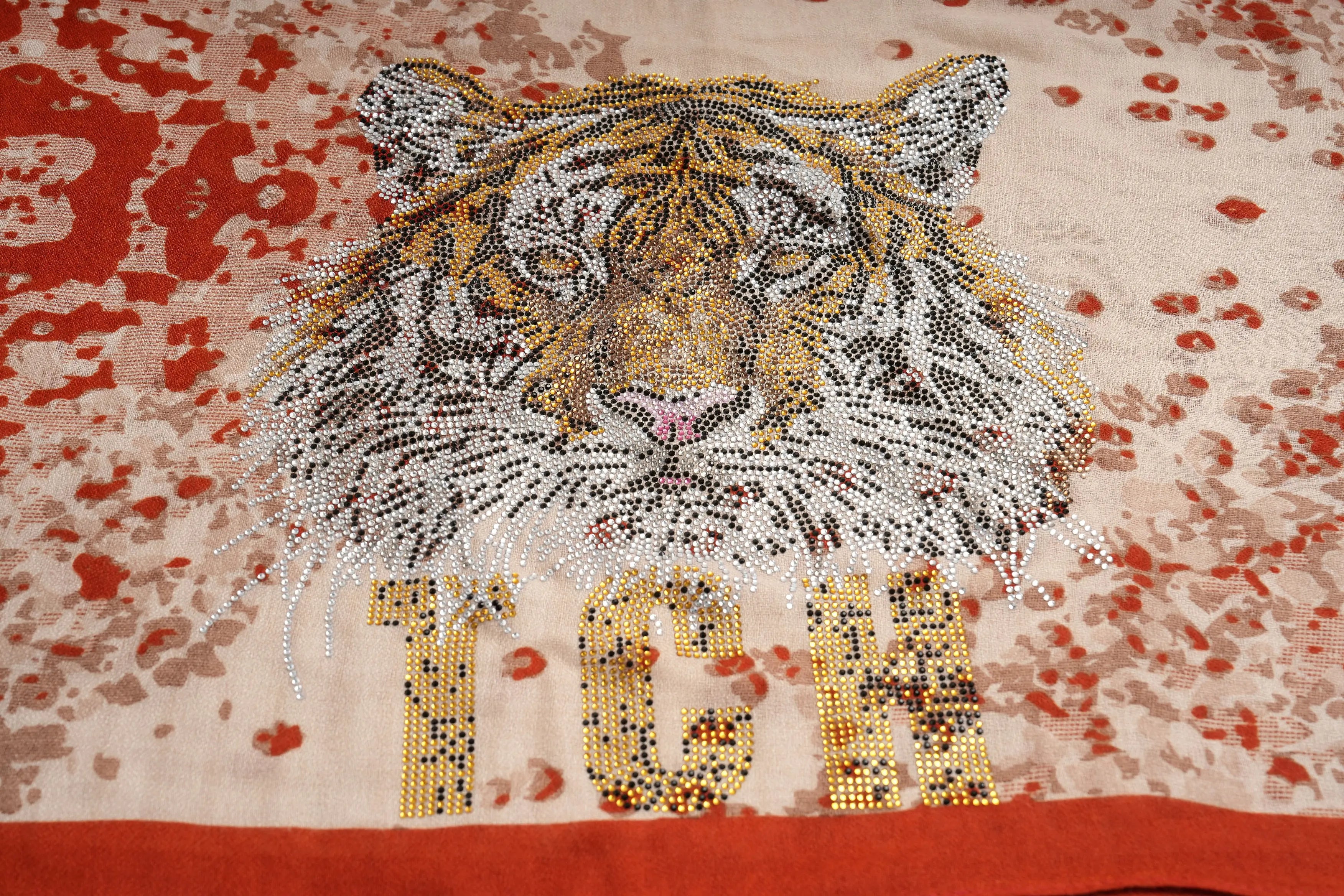 A close-up of a tiger face and the letters 'TCH' embellished with dense rhinestone beadwork on a light beige fabric with red and gold mottled patterning. TCH Vintage Leopard Print Lightweight Fashion Scarf