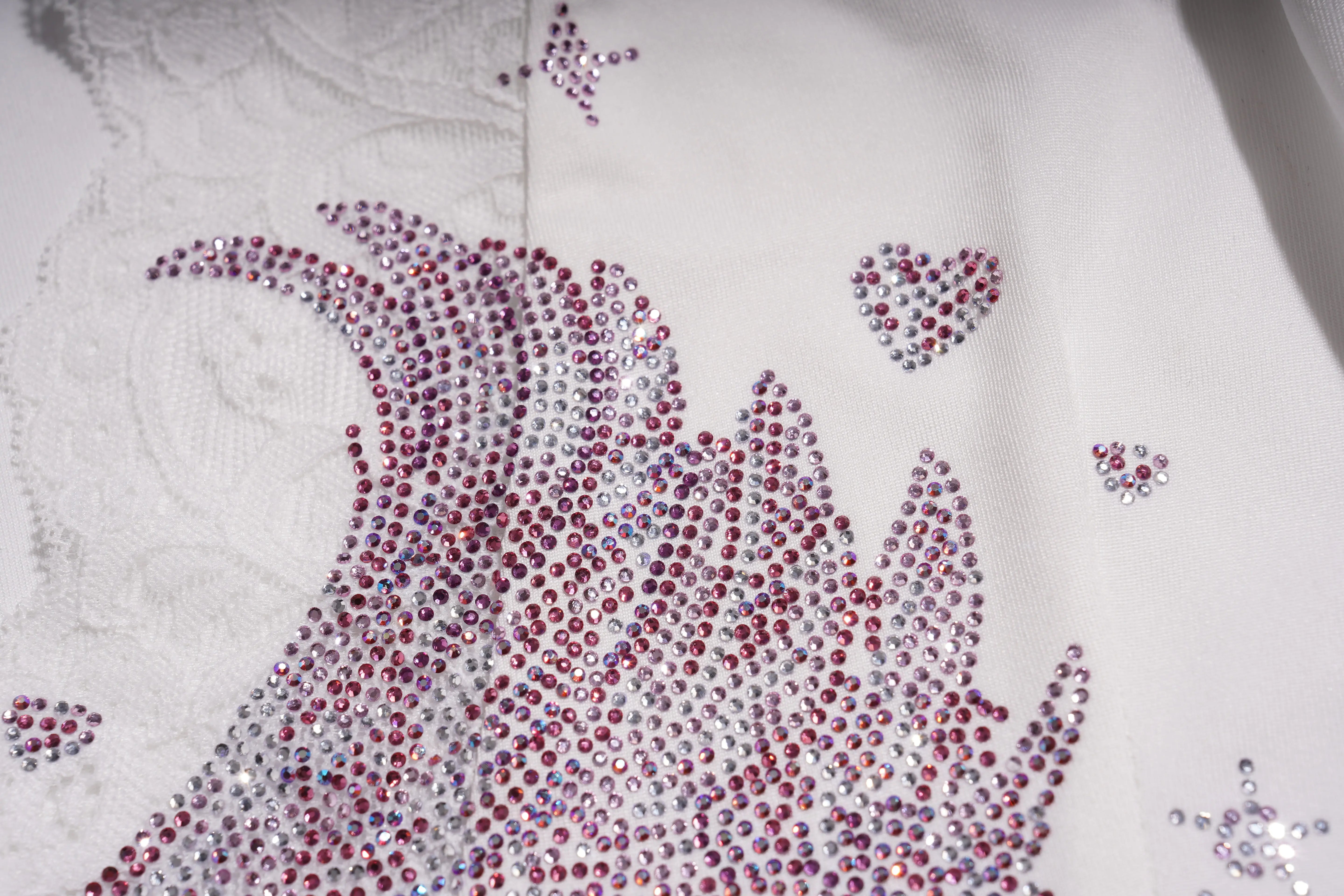 Close-up of a decorative pattern made with red and clear rhinestones on a white fabric background. TCH White Lace Trim V-Neck Rhinestone Wing Long Sleeve Top