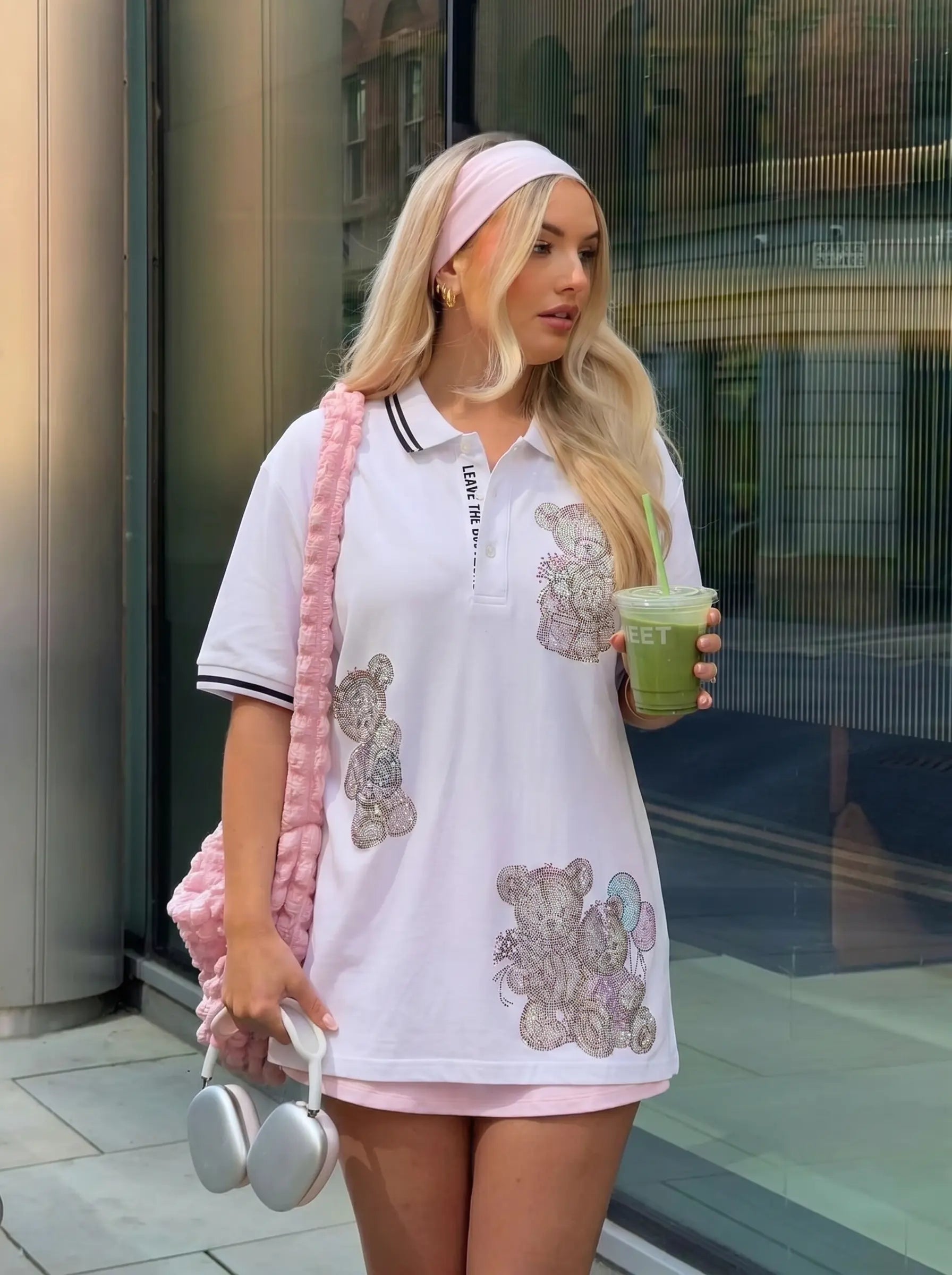 A woman wearing a white short-sleeve polo shirt with pink teddy bear prints and a pink knit shoulder bag, standing outdoors near a glass building. TCH White Teddy Bear Embellished Polo Shirt