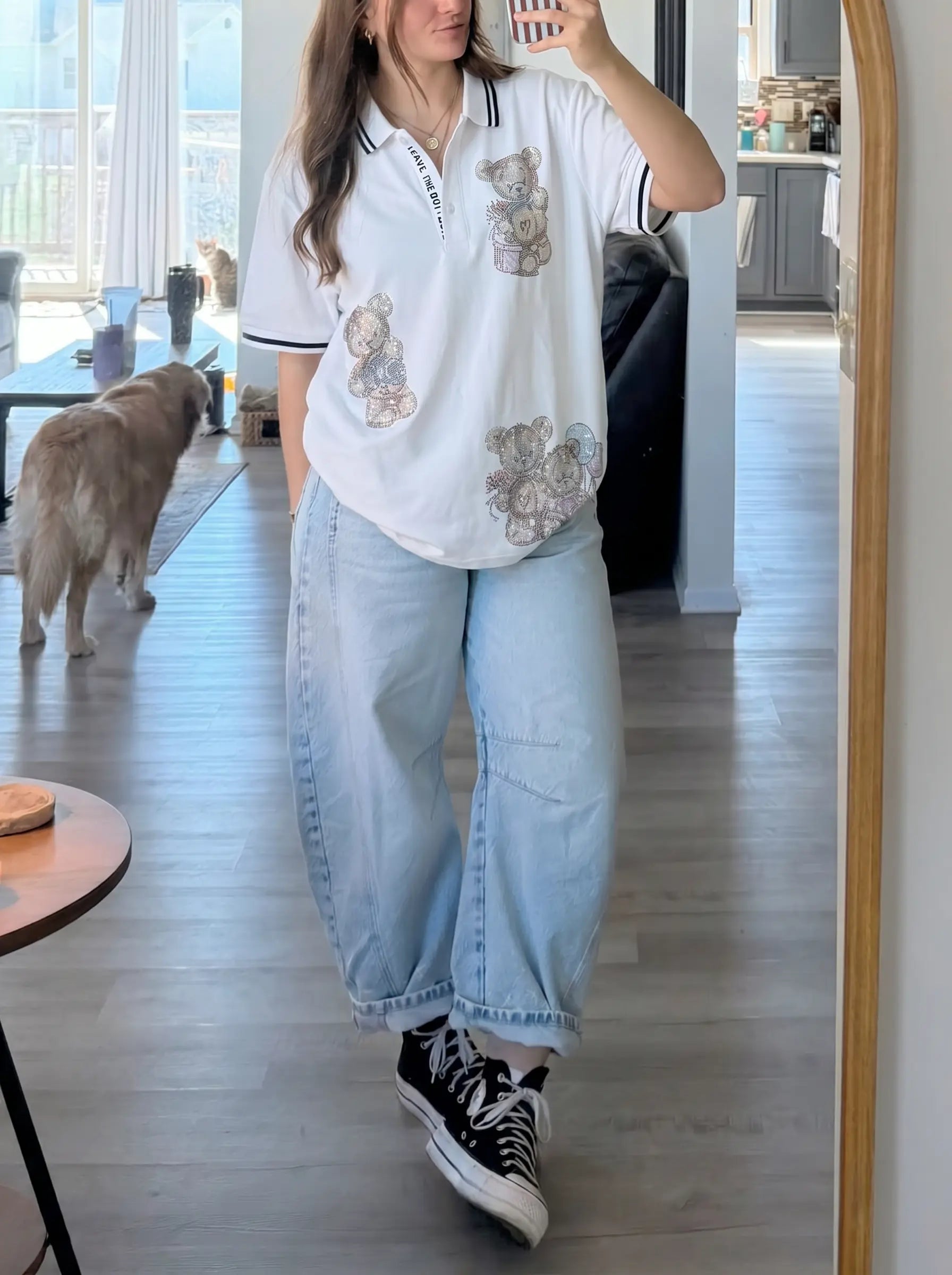 A woman takes a mirror selfie in a home setting, wearing a white short-sleeve polo shirt with embroidered teddy bear motifs, light-wash wide-leg jeans, and black low-top sneakers. A dog is visible in the background near a window. TCH White Teddy Bear Embellished Polo Shirt